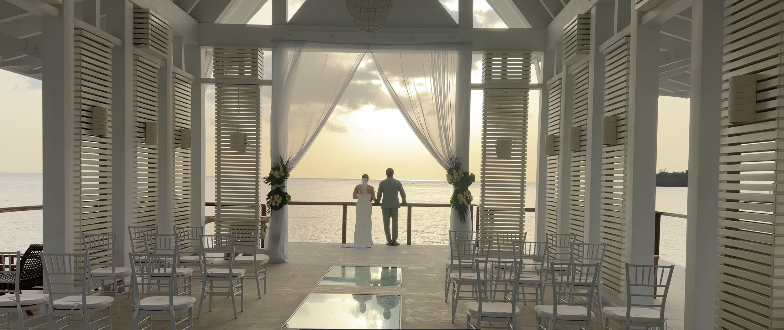 A couple standing on a balcony overlooking the ocean during sunset at a wedding ceremony venue decorated with drapery and floral arrangements.