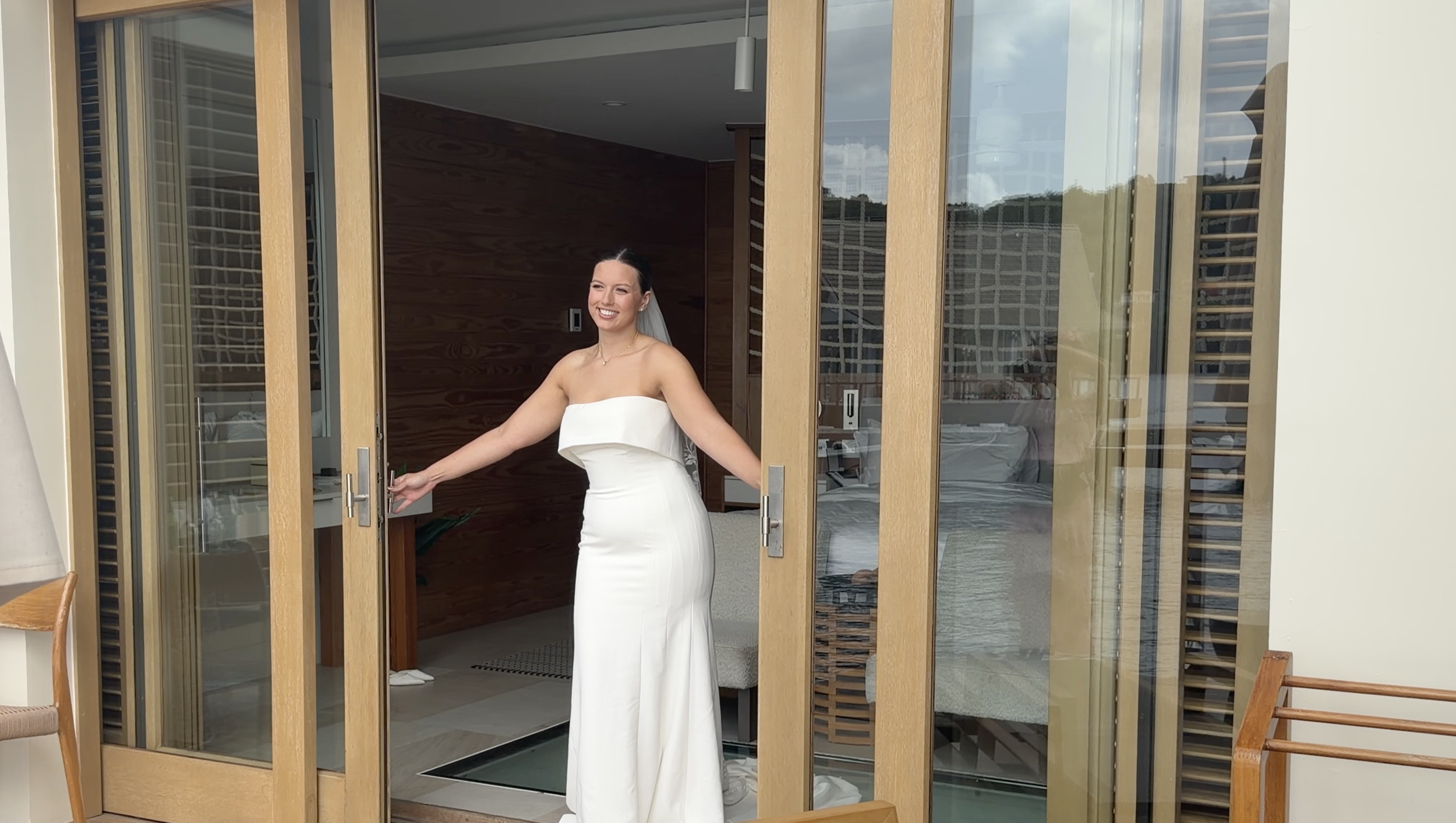 A woman in a white strapless dress standing at an open doorway, smiling, with a wooden wall inside and a bed visible through the glass doors.