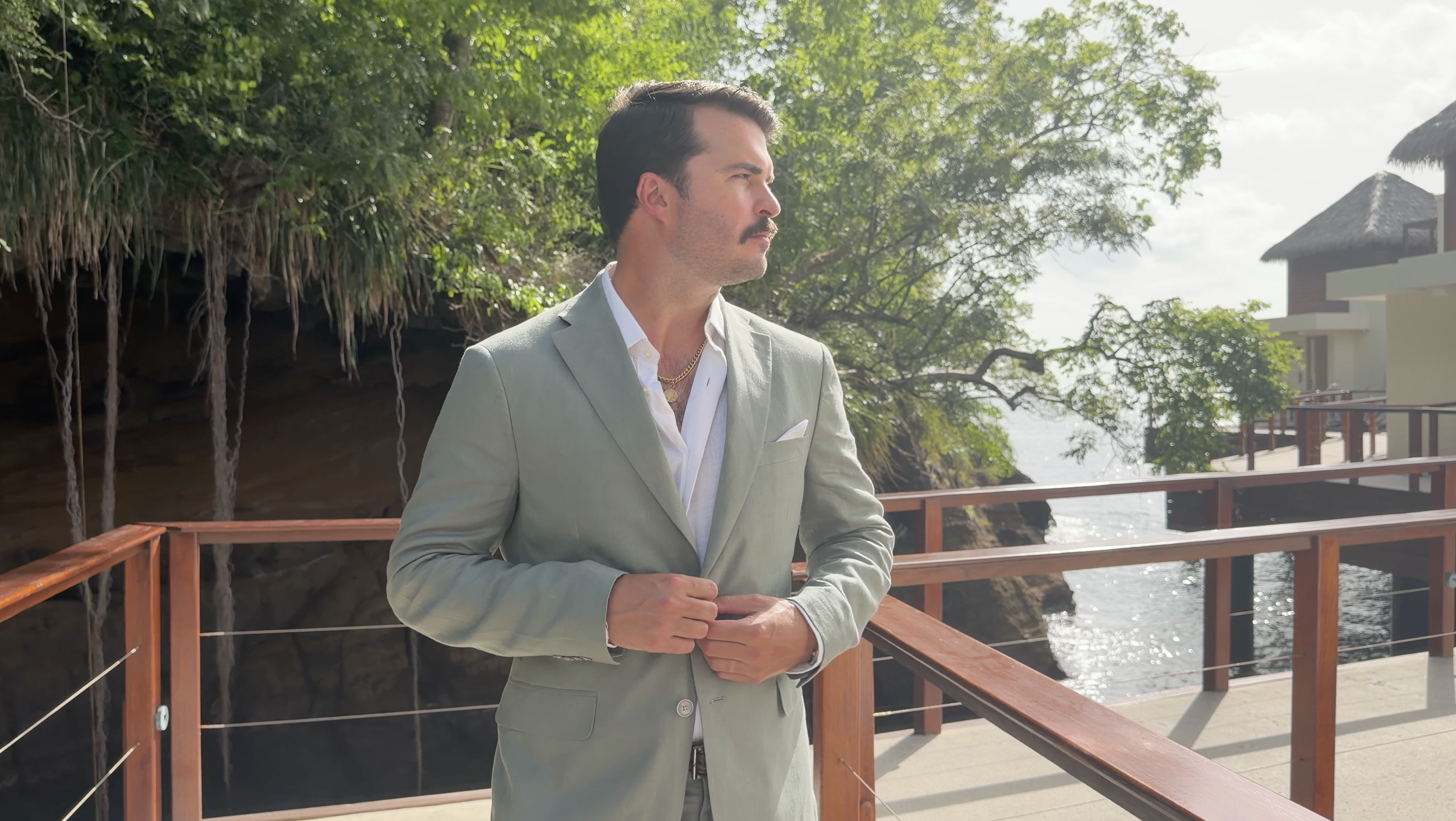 A man in a light gray suit with a white shirt and gold chains standing on a wooden deck by the water, with green trees and buildings in the background.