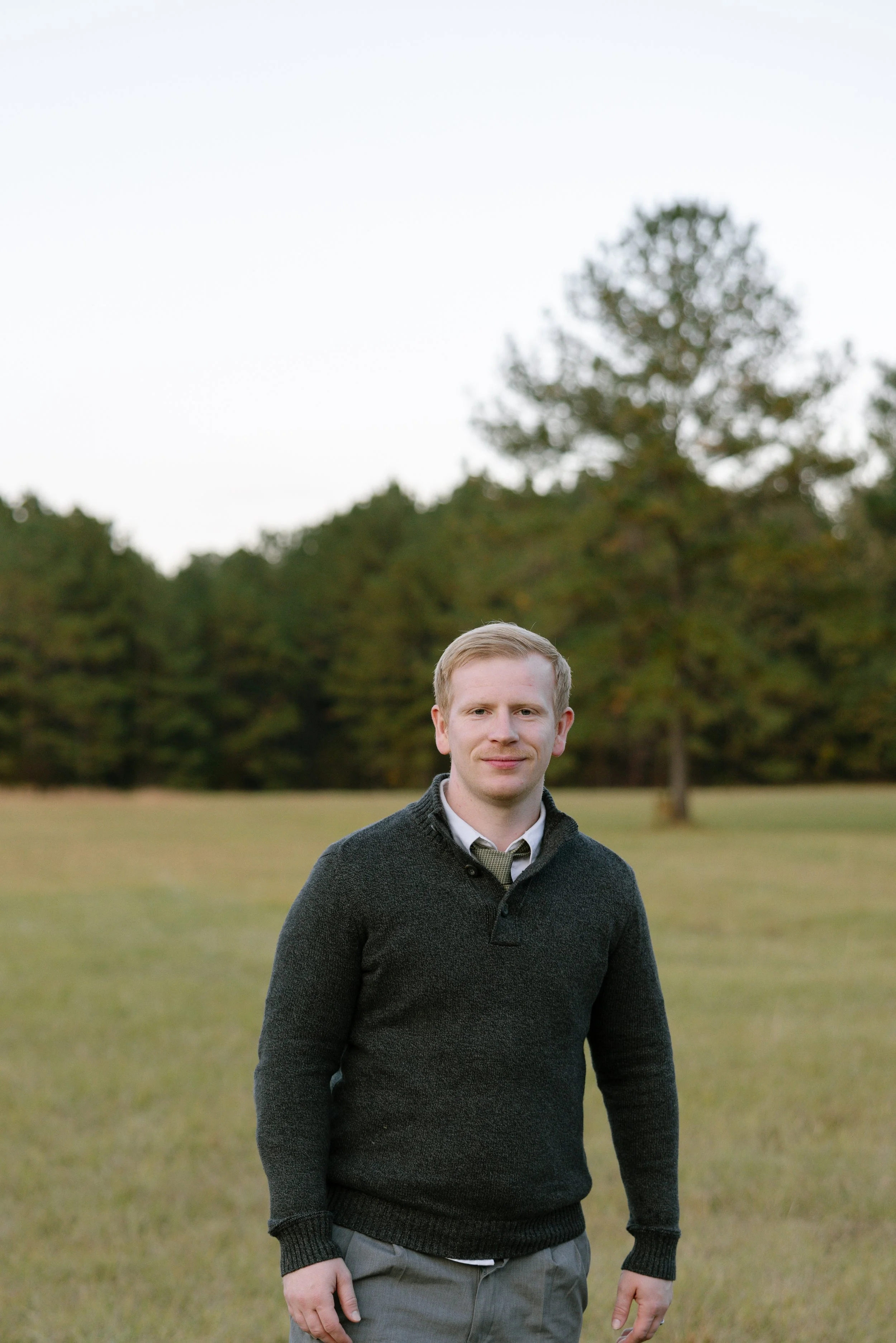 Men wearing a dark sweater and gray pants standing in an open grassy field with trees in the background under an overcast sky.