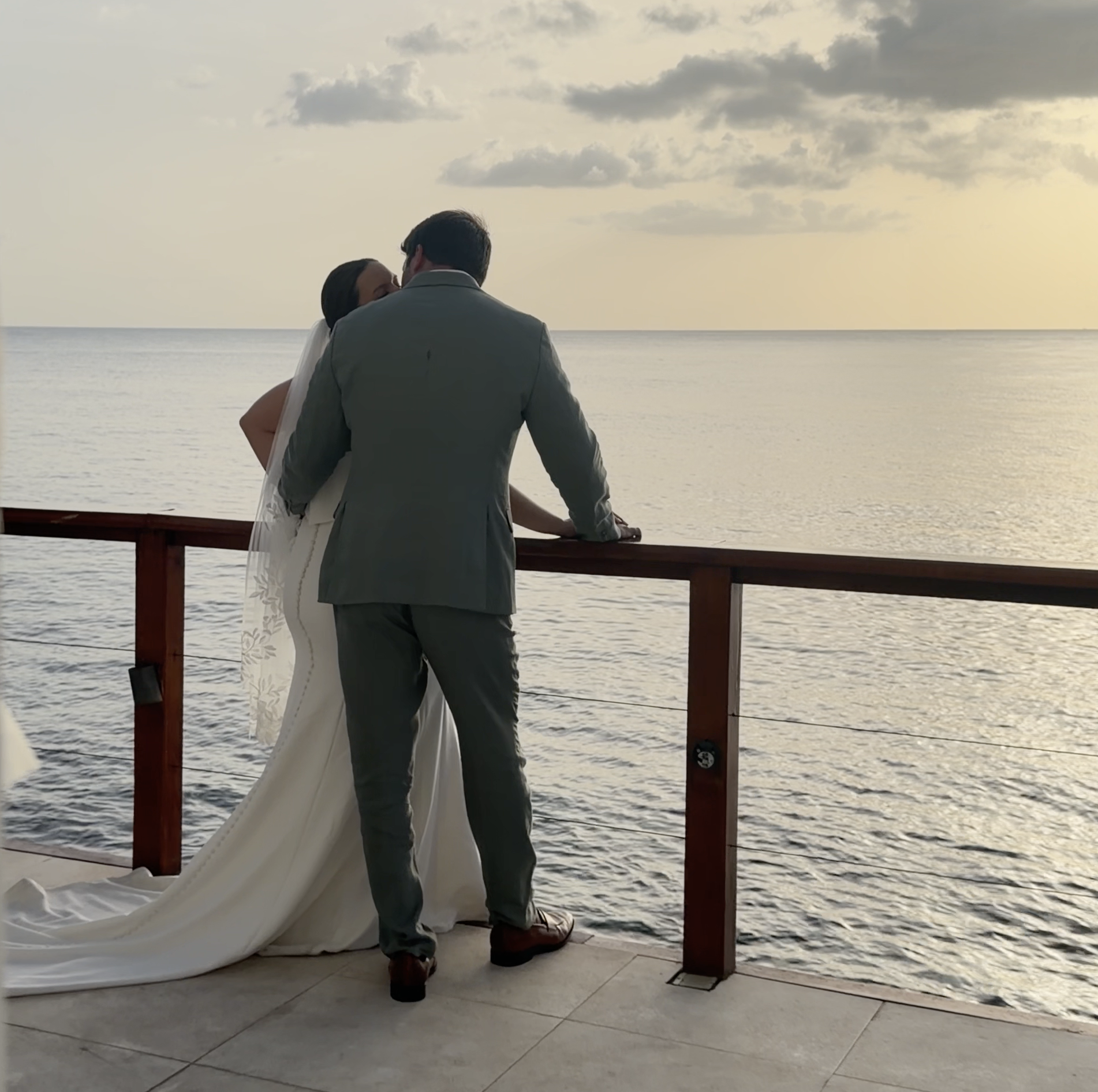 A couple dressed in wedding attire standing on a balcony overlooking the ocean during sunset, sharing an intimate moment