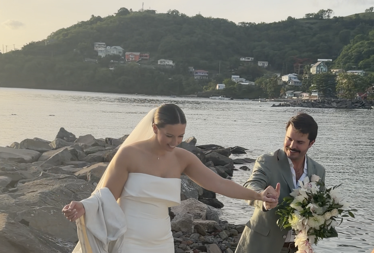 A bride and groom holding hands on a rocky riverside during their wedding ceremony, with houses on a hillside in the background.