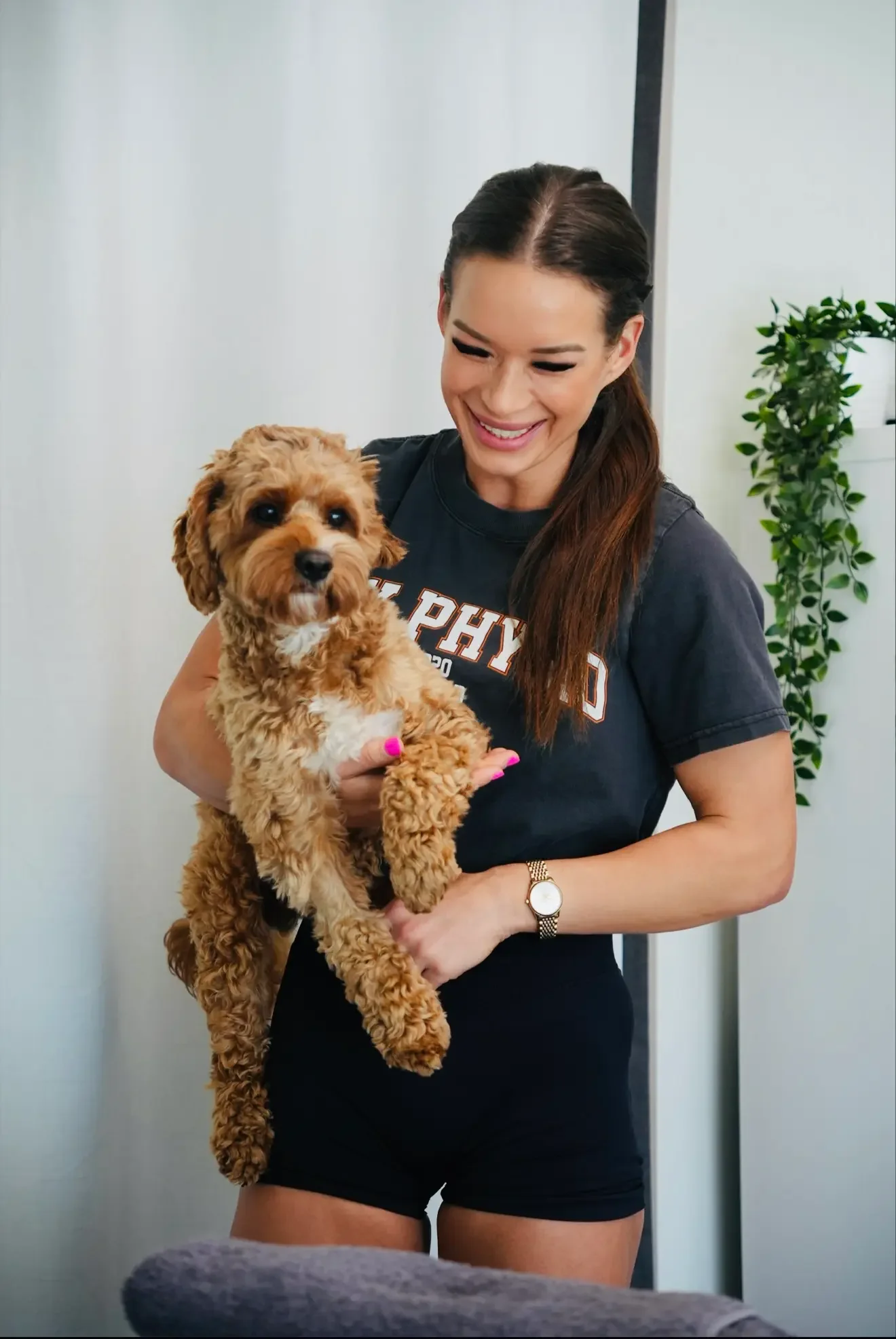 A woman with long brown hair is smiling while holding a small brown curly-haired dog with white markings. The woman is wearing a dark T-shirt and black shorts. She is standing indoors near a white wall and a green potted plant.