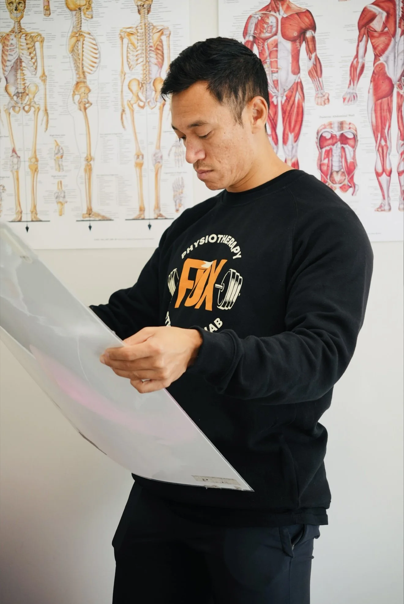 A man in a Giants hoodie looking at a chart or paper in a medical or anatomical setting with anatomical posters of muscles and skeletons in the background.