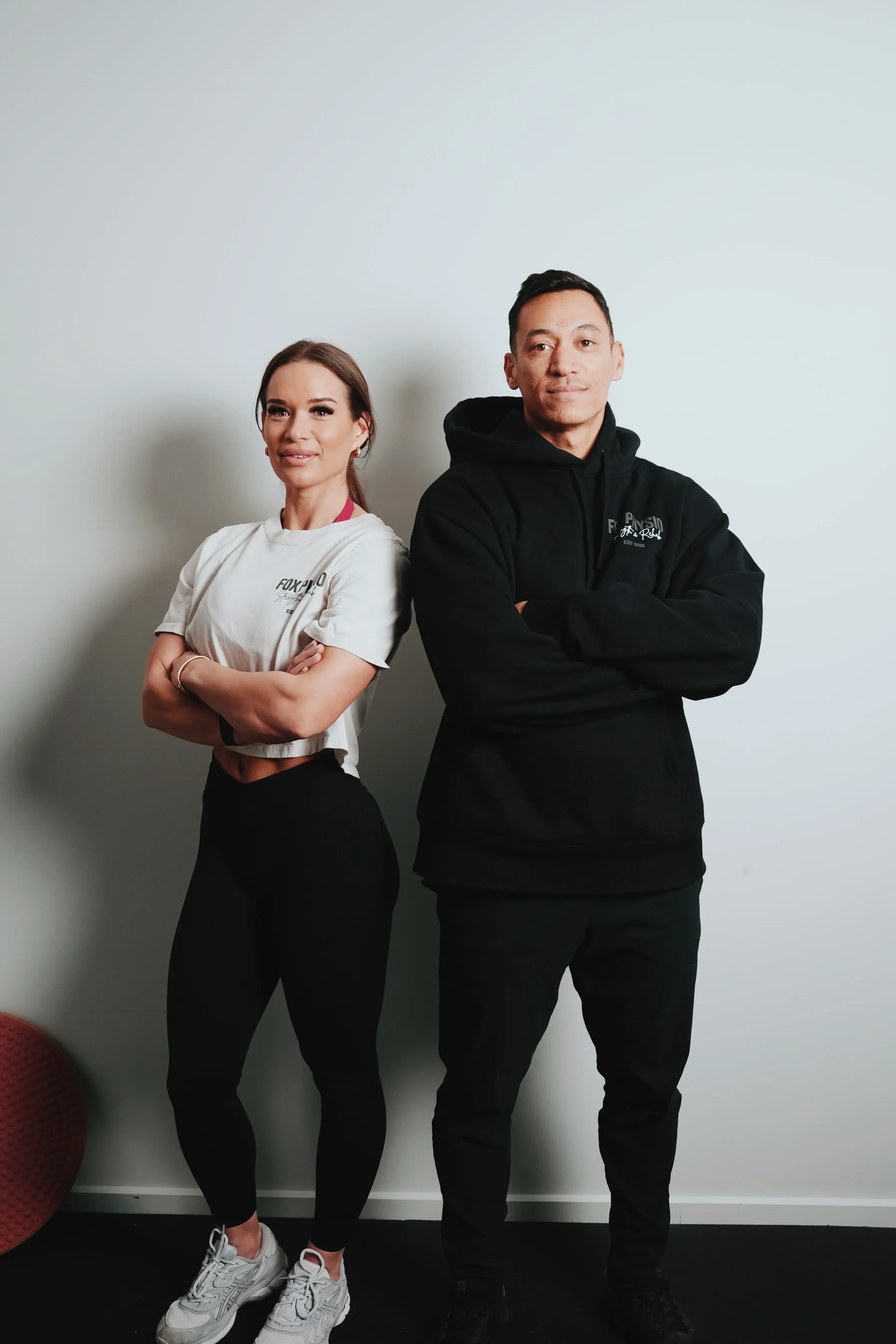 A woman with long brown hair, wearing a white t-shirt and black pants, standing with her arms crossed, smiling. A man with short black hair, wearing a black hoodie and black pants, also standing with his arms crossed, looking at the camera. They are standing against a plain light-colored wall.