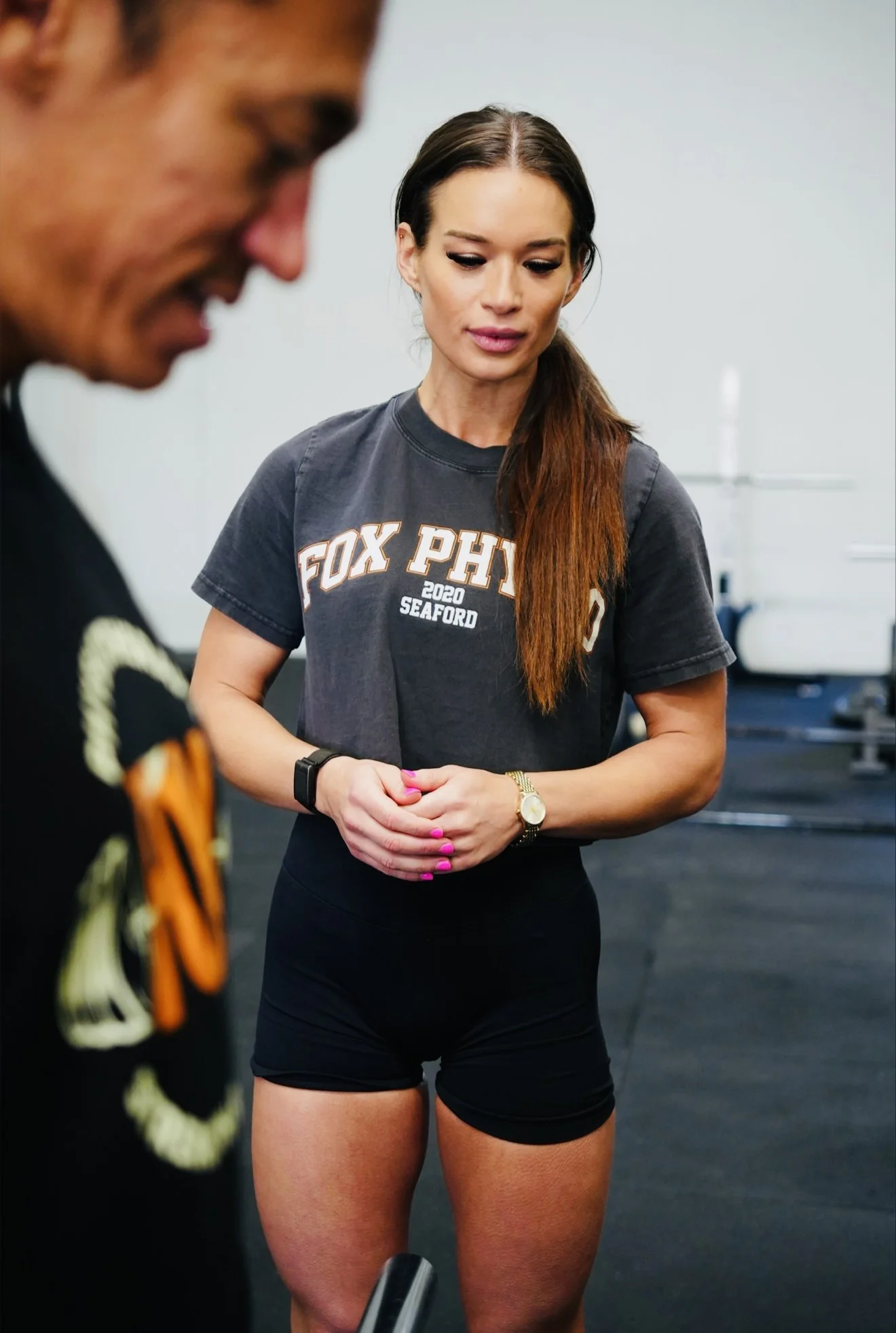 A woman in athletic clothing, including a gray T-shirt and black shorts, standing in a gym with her hands clasped. Partially visible person in foreground appears to be in a costume or uniform.