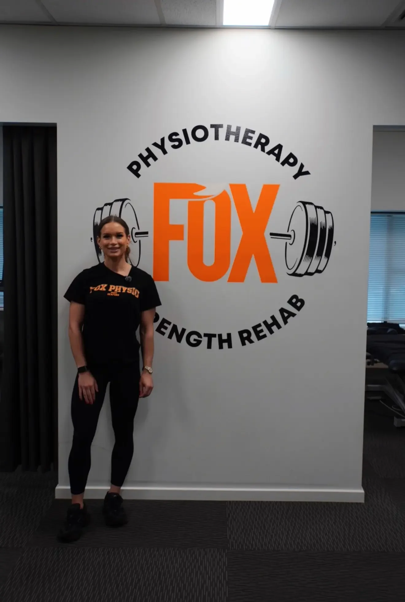 A woman in black workout clothes standing in front of a wall with a logo that says 'Physiotherapy FUX Strength Rehab' and an illustration of a barbell.