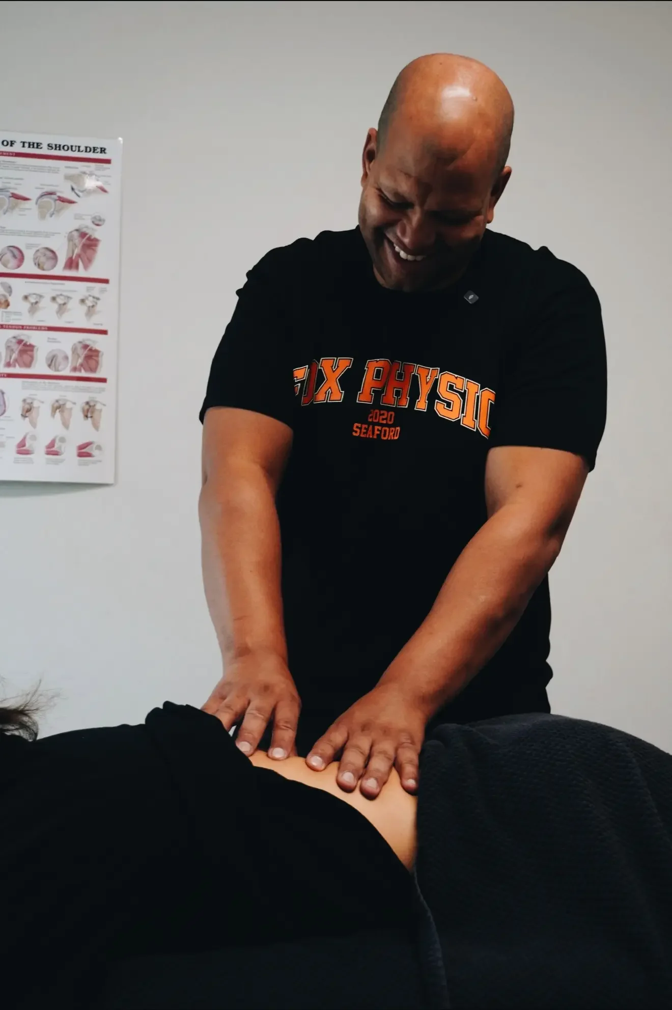 A person performing a physical therapy or chiropractic treatment, pressing on a patient's abdomen while smiling. The setting appears to be a clinic with a poster about shoulder anatomy on the wall.