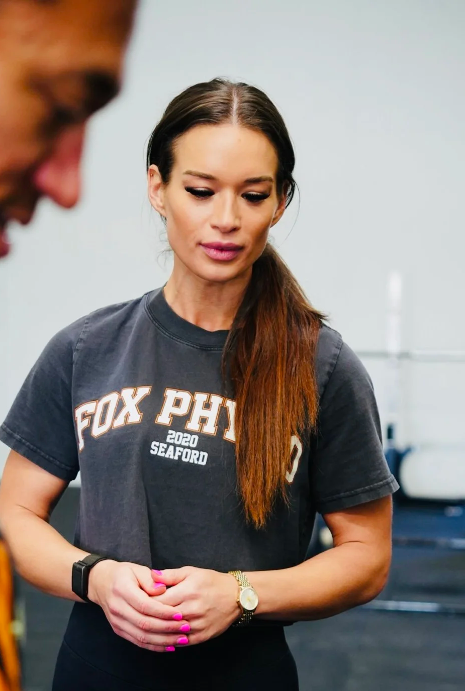 A woman with long reddish-brown hair, wearing a dark gray FOX PHX 2020 Seaford T-shirt, looking downward with hands clasped, standing in an indoor setting.