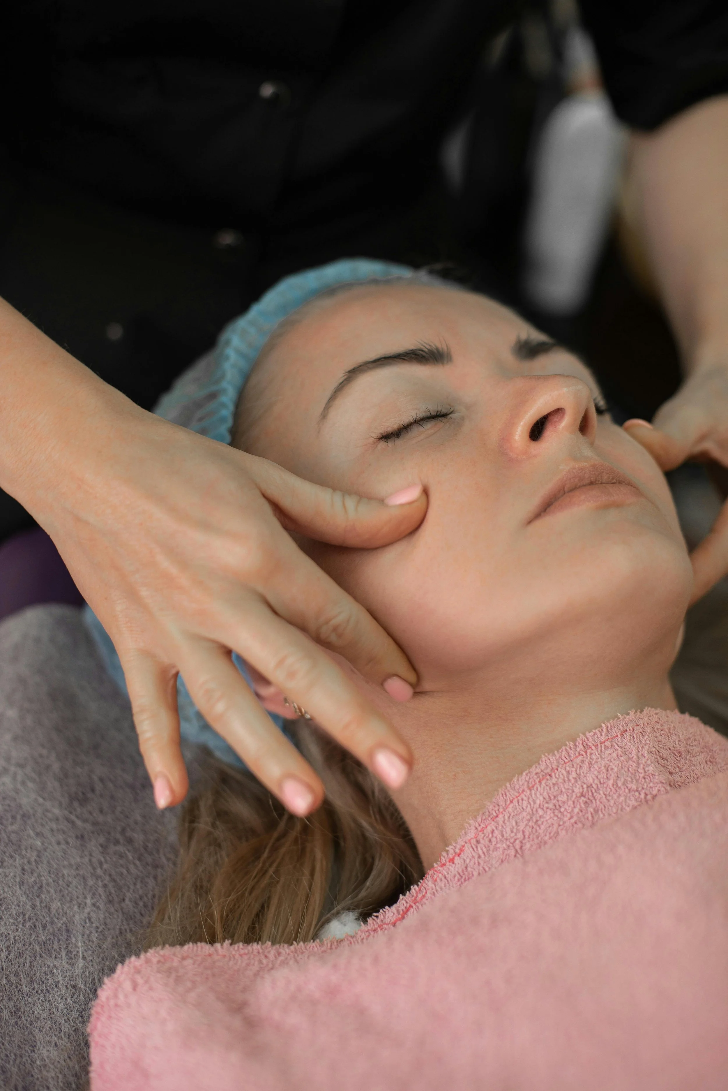 A woman receiving a facial massage while lying down with eyes closed, wearing a pink towel and a blue head covering.