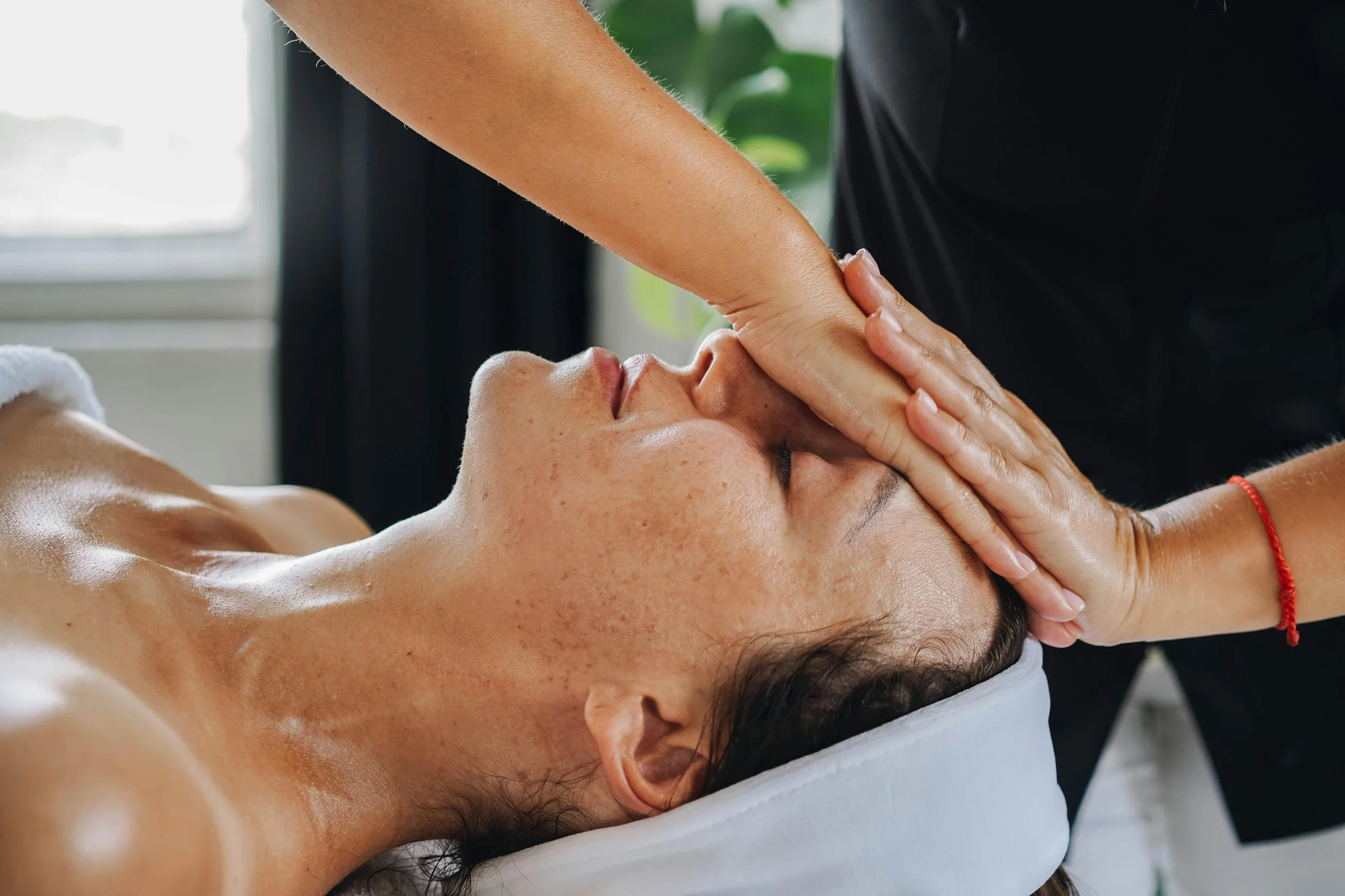 A person receiving a facial massage or treatment, lying down with eyes closed, while a professional performs a facial massage on their face in a bright, relaxing setting.