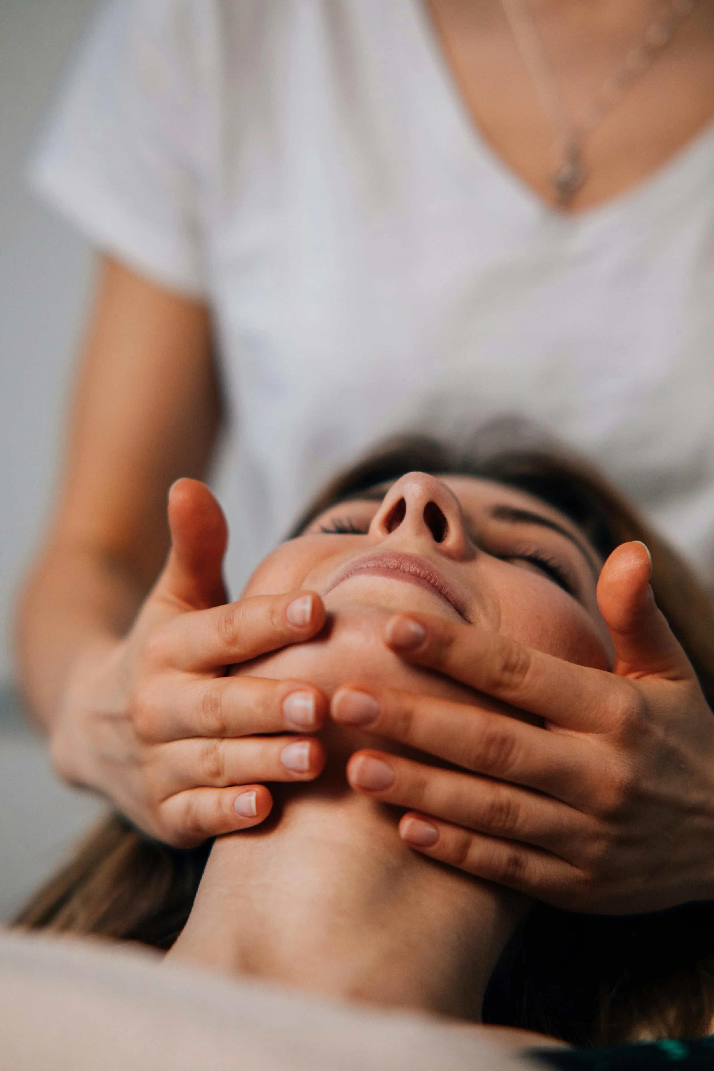 Close-up of a person lying down, with their eyes closed, receiving a facial treatment from a practitioner. The practitioner is gently holding the person's face with both hands.