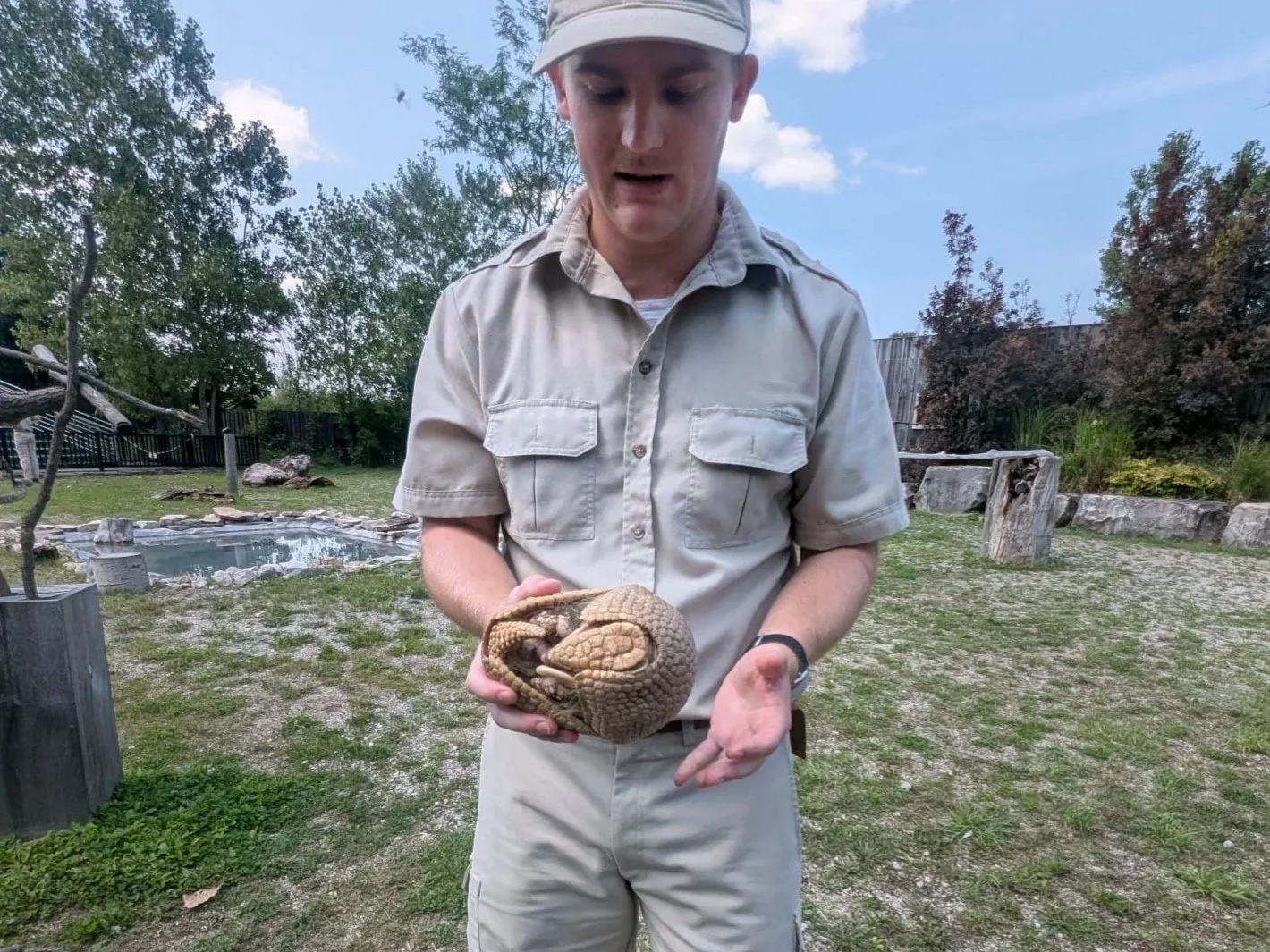A young man wearing a beige safari shirt and hat holding a curled-up, small, furry animal in his hands outdoors with trees, a fence, and a small pond in the background.