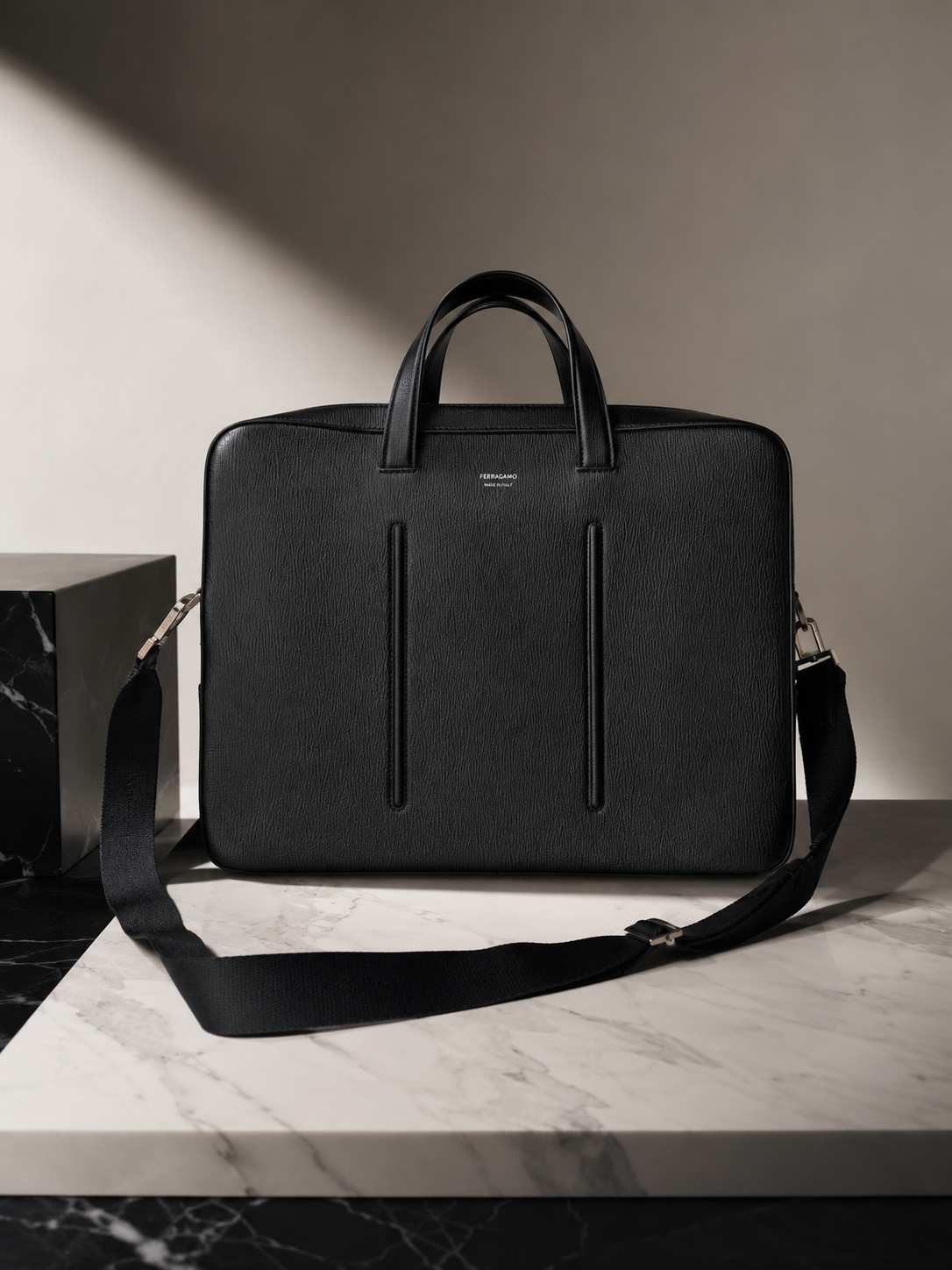 Maletín negro de cuero con asas y correa ajustable, sobre mesa de mármol blanco y negro.