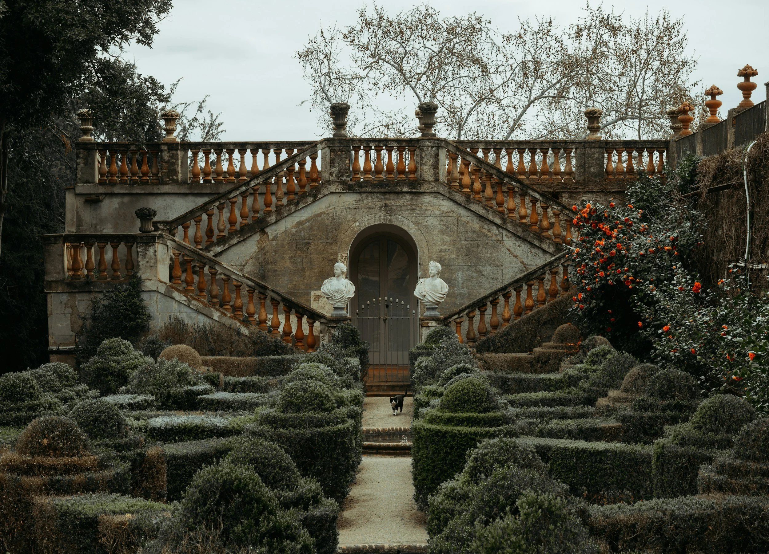 Escalera en un jardín antiguo con estatuas de bustos y macetas de cerámica, rodeada de plantas y flores, con árboles sin hojas al fondo y un perro pequeño en el sendero.