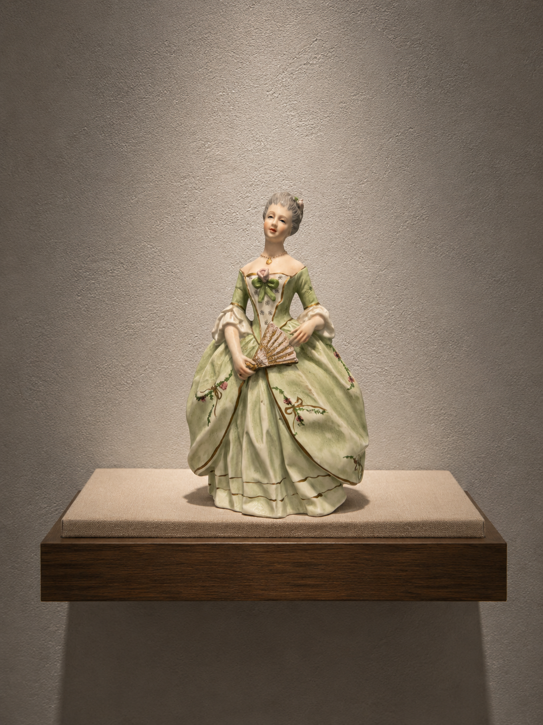 Escultura de una mujer de estilo rococó con vestido verde y detalles florales, sosteniendo un abanico, exhibida en una galería de arte.