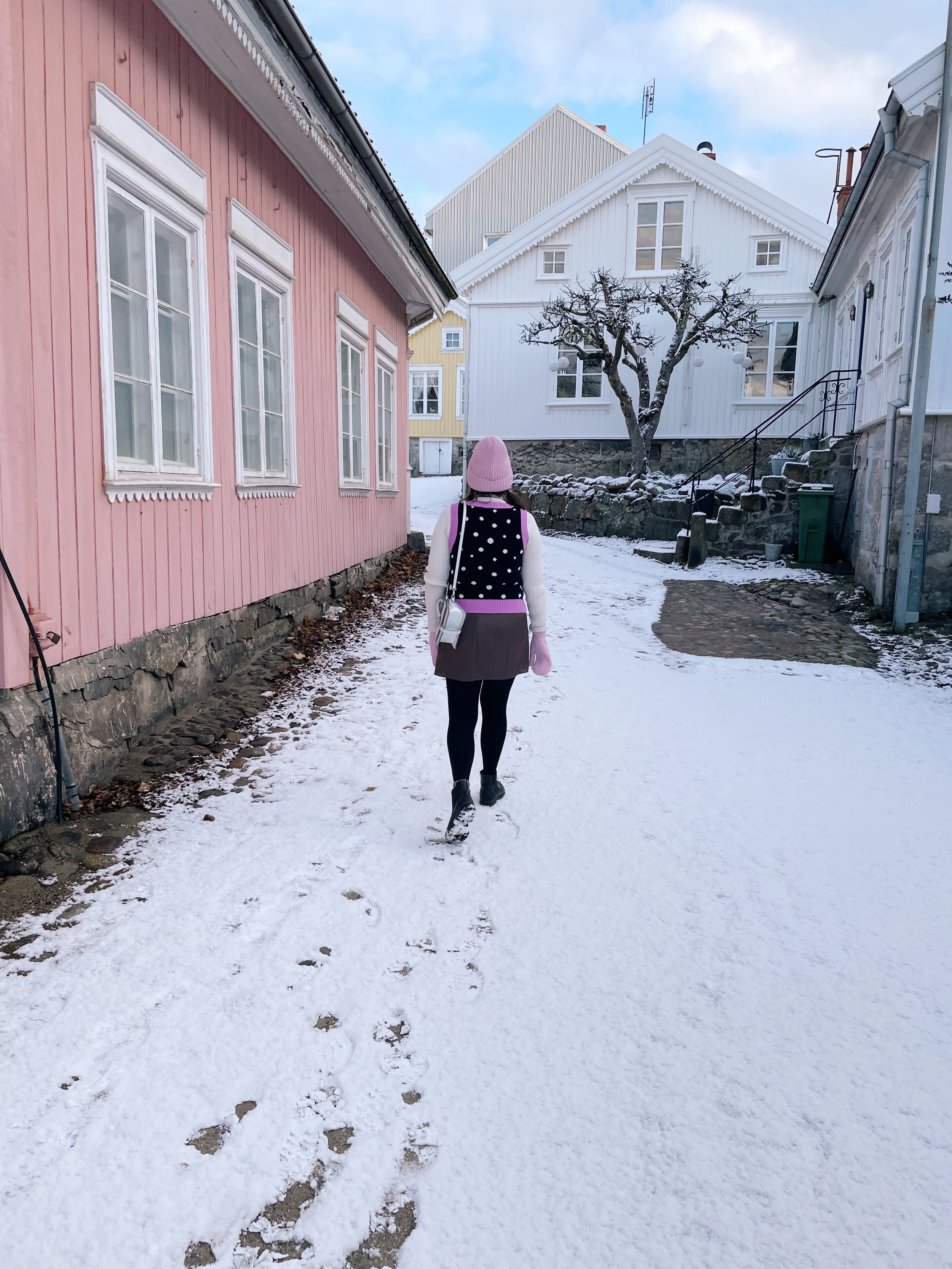 A person walking on a snow-covered path between colorful houses, wearing a pink beanie, pink gloves, a black and white polka dot vest, and black pants, with footprints visible in the snow.