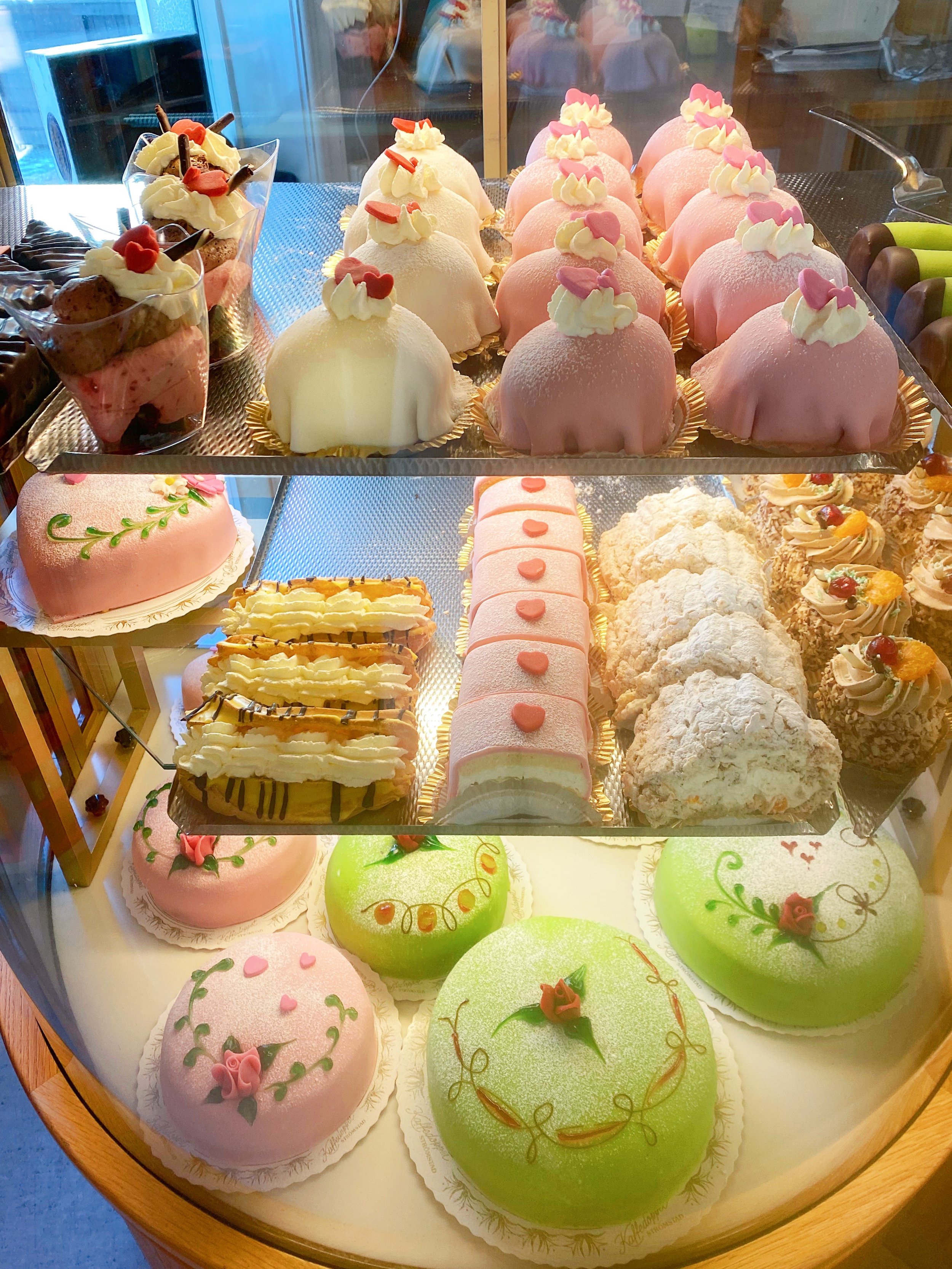 A display case filled with various decorated cakes and pastries, including pink and white dome-shaped desserts topped with whipped cream and hearts, oval cakes with floral decorations, and other intricately decorated confections.