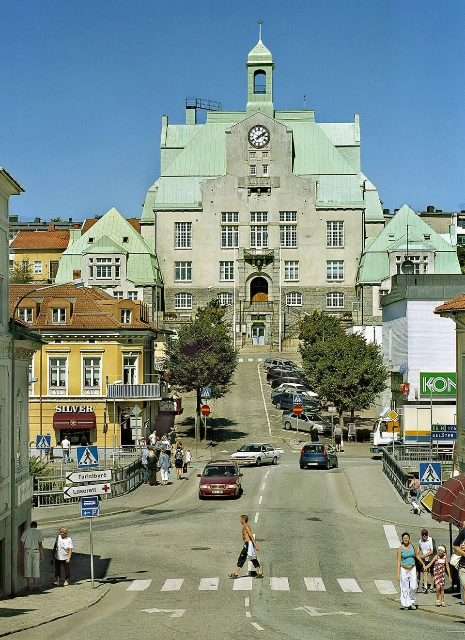 Street scene with pedestrians crossing, parked cars, and a large historic building with a green copper roof and a clock on the facade in the background.