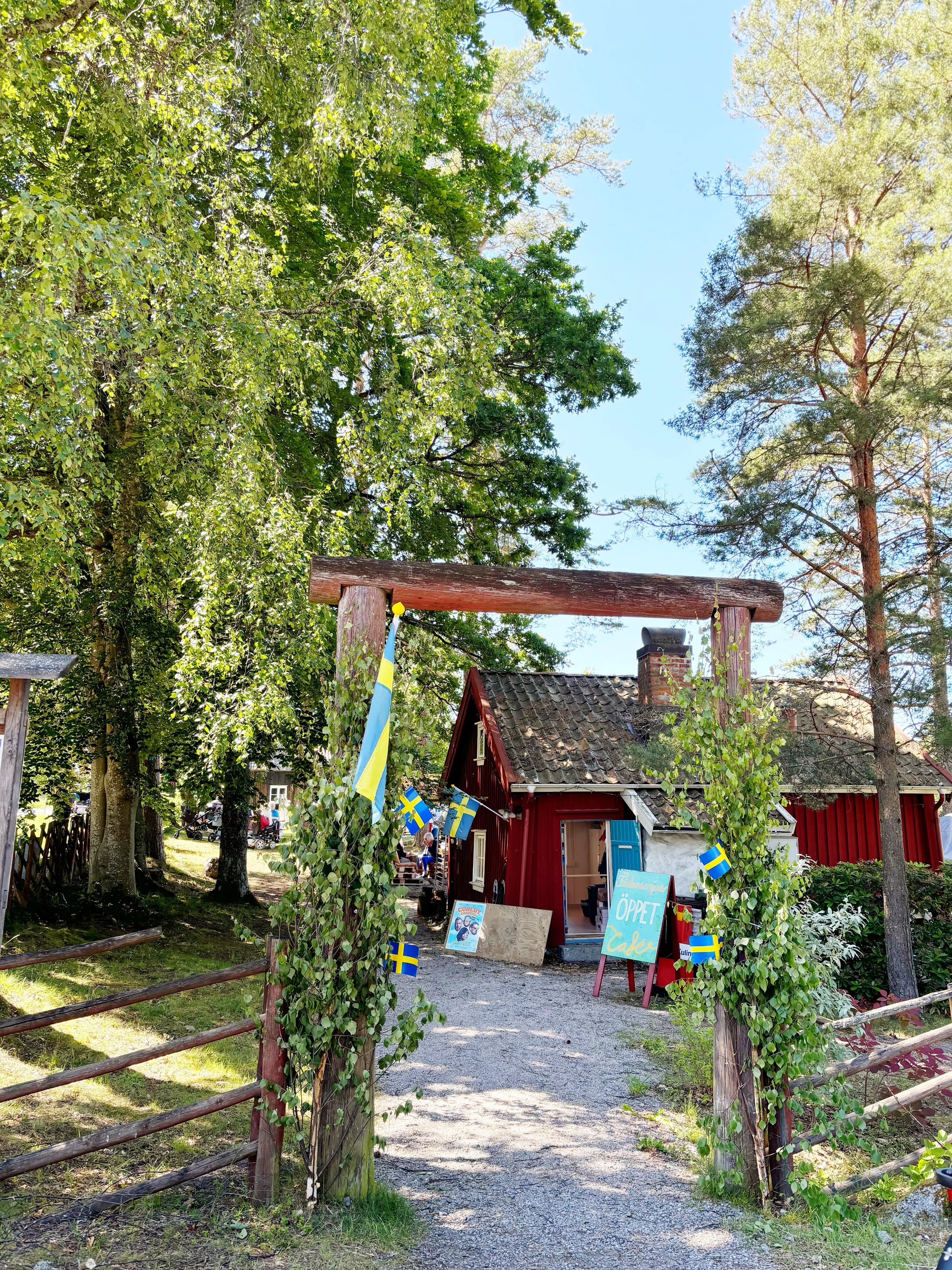 A red wooden building decorated with small Swedish flags. The building is surrounded by green trees and bushes with a bright blue sky overhead. A sign in front of the building says 'Öppet' and 'Café'. There are small outdoor seating areas and people in the background.