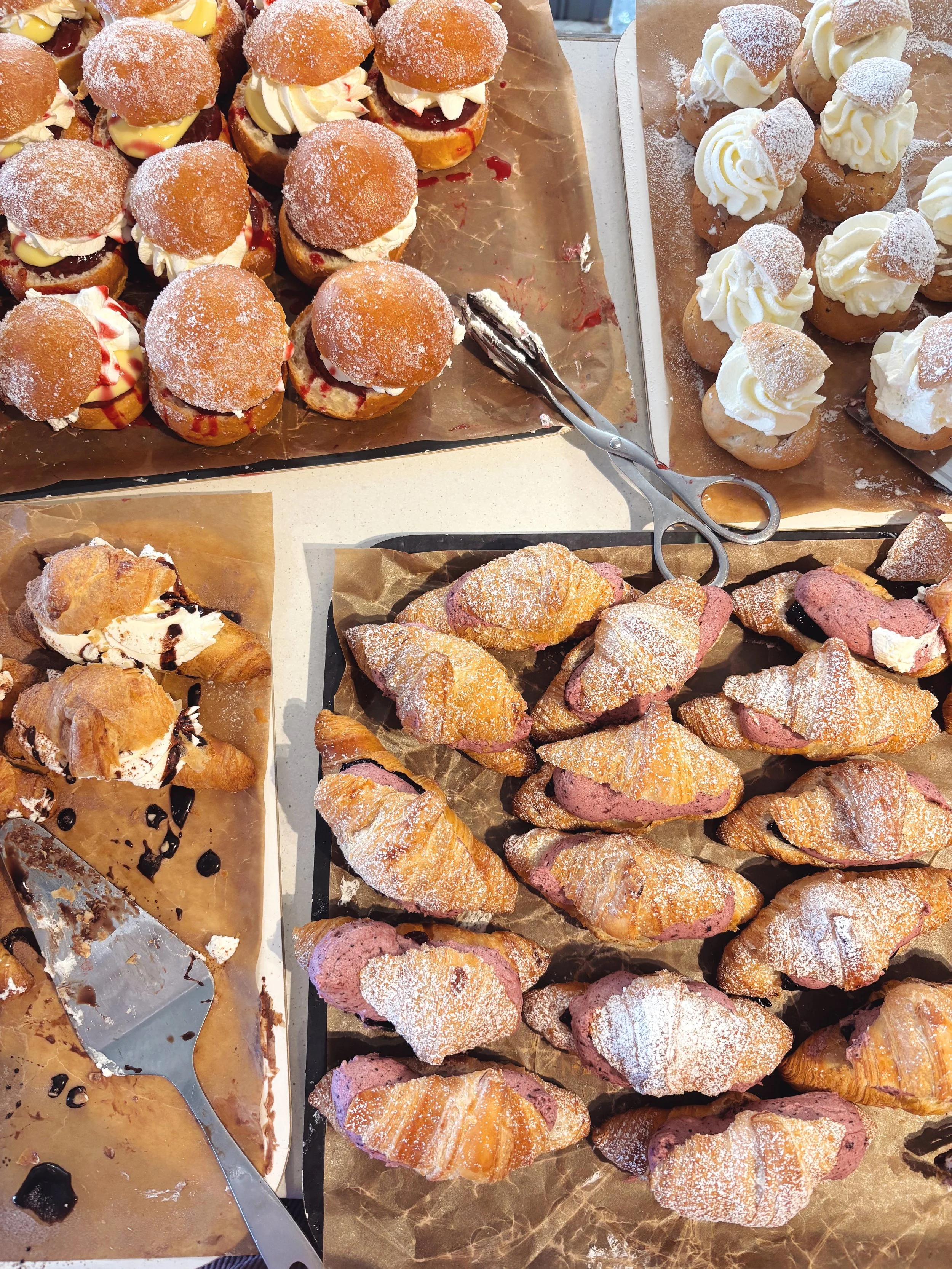 Display of various bakery pastries including cream-filled croissants, donuts topped with powdered sugar, and fruit-filled treats on parchment paper