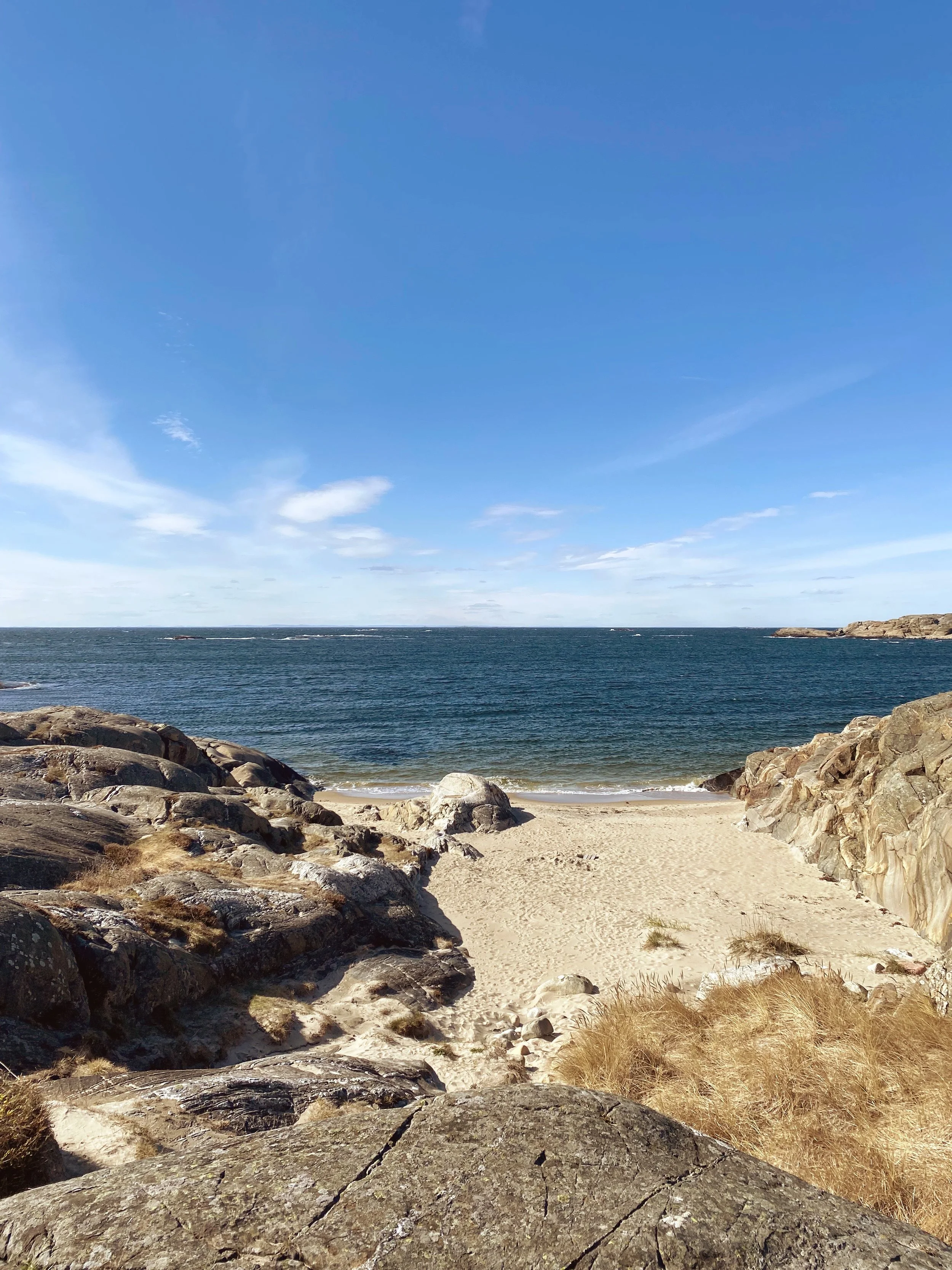 Beach scene with sandy shore, large rocks in the foreground, and a calm ocean under a partly cloudy sky.