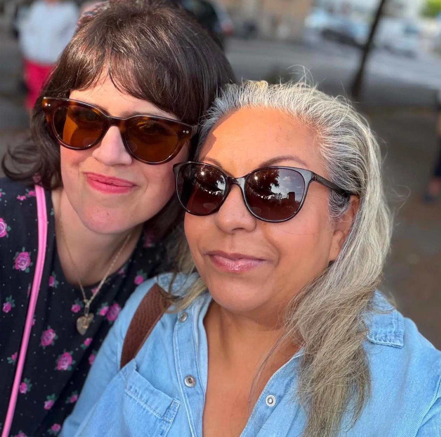 Two women wearing sunglasses taking a selfie outdoors, with trees and cars in the background.