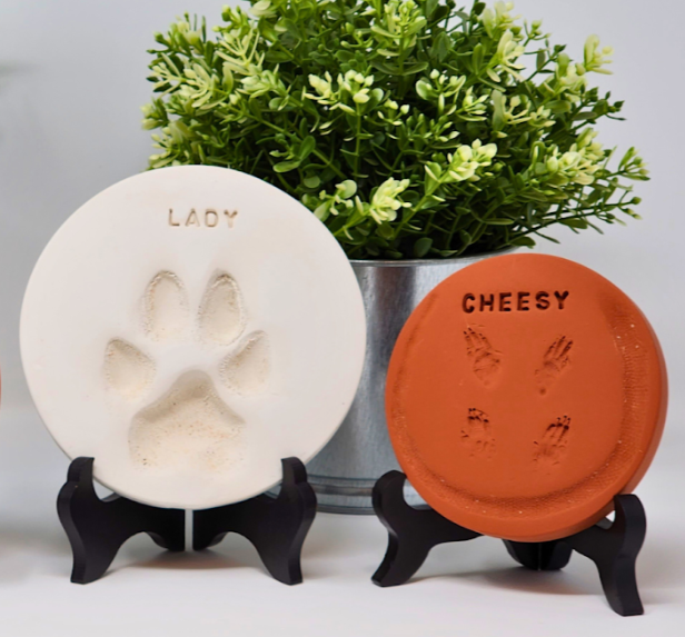 Two pet-themed soap bars, one shaped like a paw print labeled "Lady" in white, and the other shaped like an animal face labeled "Cheesy" in brown, positioned on small black stands in front of a potted green plant.