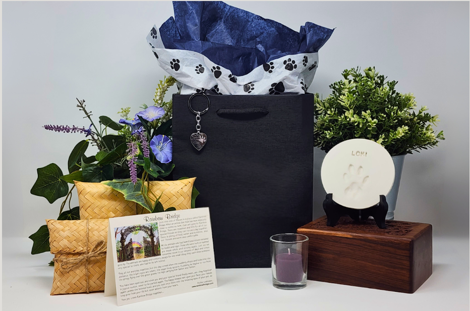 Arrangement of gift and decorative items including a black gift bag, potted green plants with purple flowers, a white soap with a paw print and the word "Loki" on a black stand, a small candle in a glass holder, and a printed card with information about a rainbow bridge, all set against a plain background.
