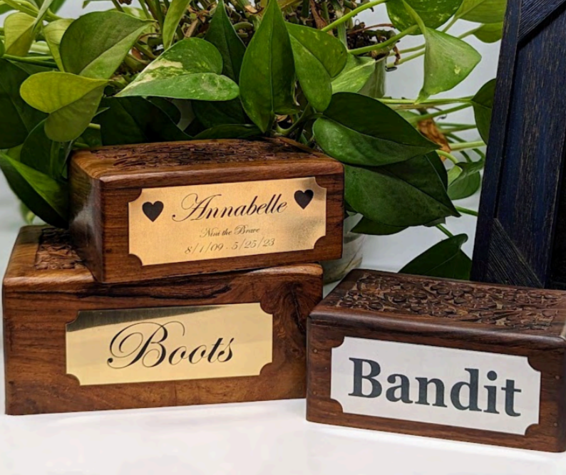 Three wooden boxes with engraved labels: one says "Annabelle" with date and birth details, another says "Boots," and the third says "Bandit." They are displayed with greenery in the background.