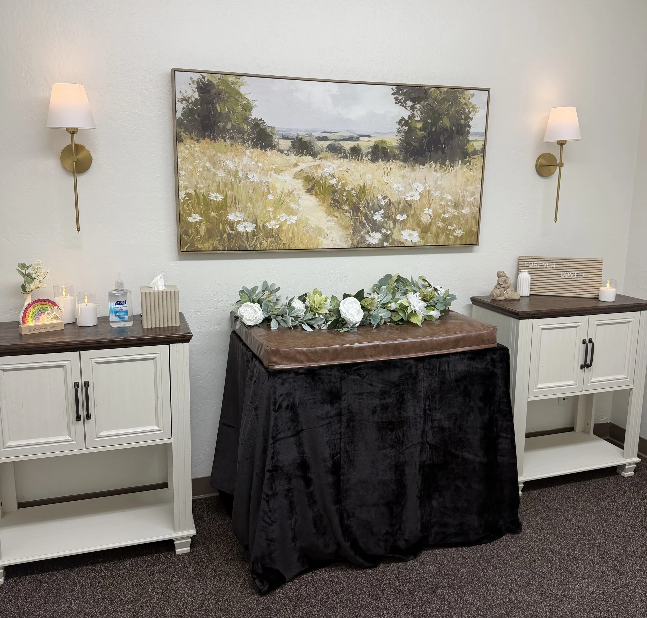 A floral arrangement with greenery and white roses on a table covered with a dark cloth, in front of a painting of a landscape with trees and a path. Two wall-mounted lamps with white shades flank the painting.