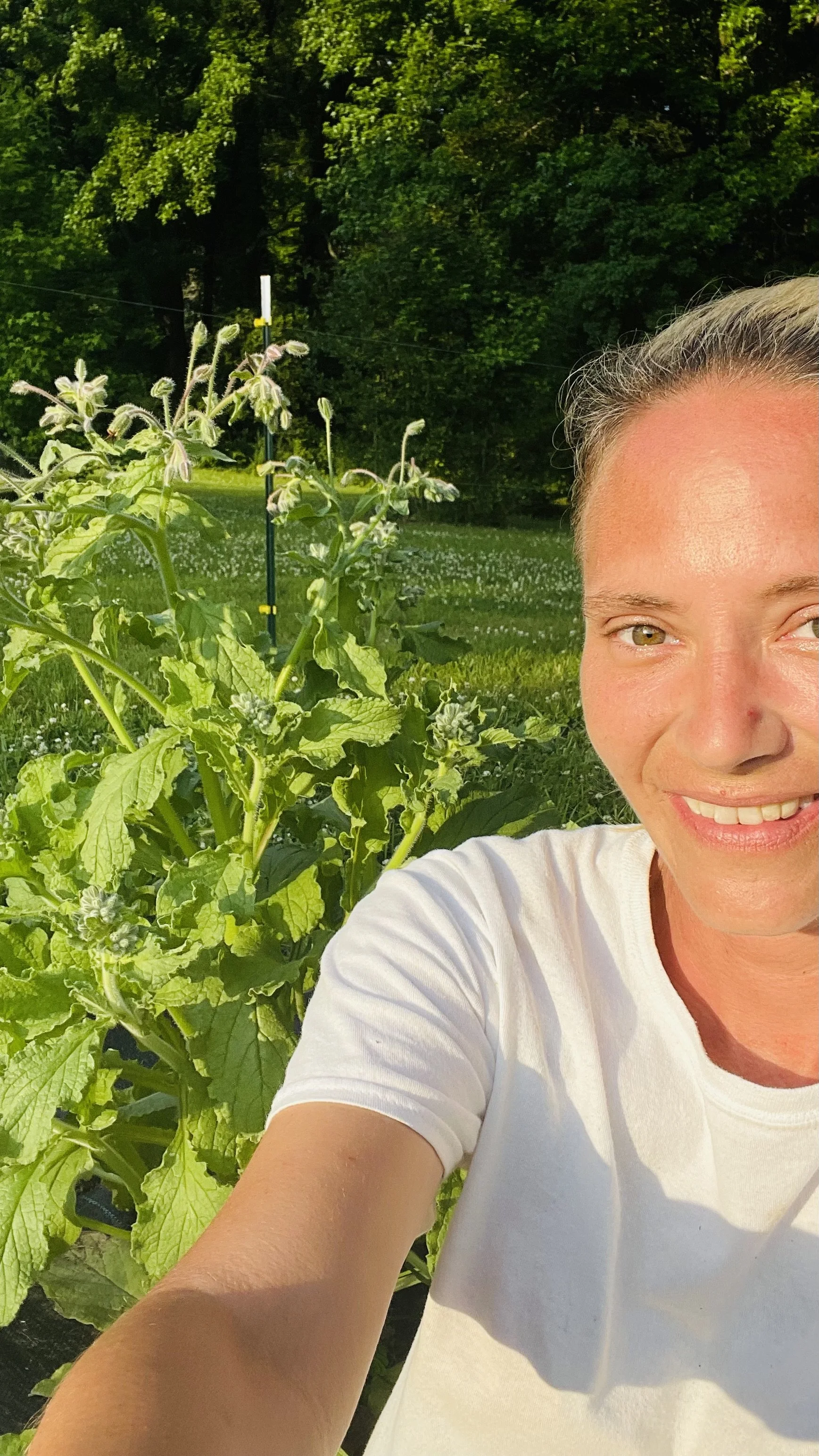 A woman taking a selfie outdoors in a garden with green plants and trees in the background, smiling and wearing a white t-shirt.