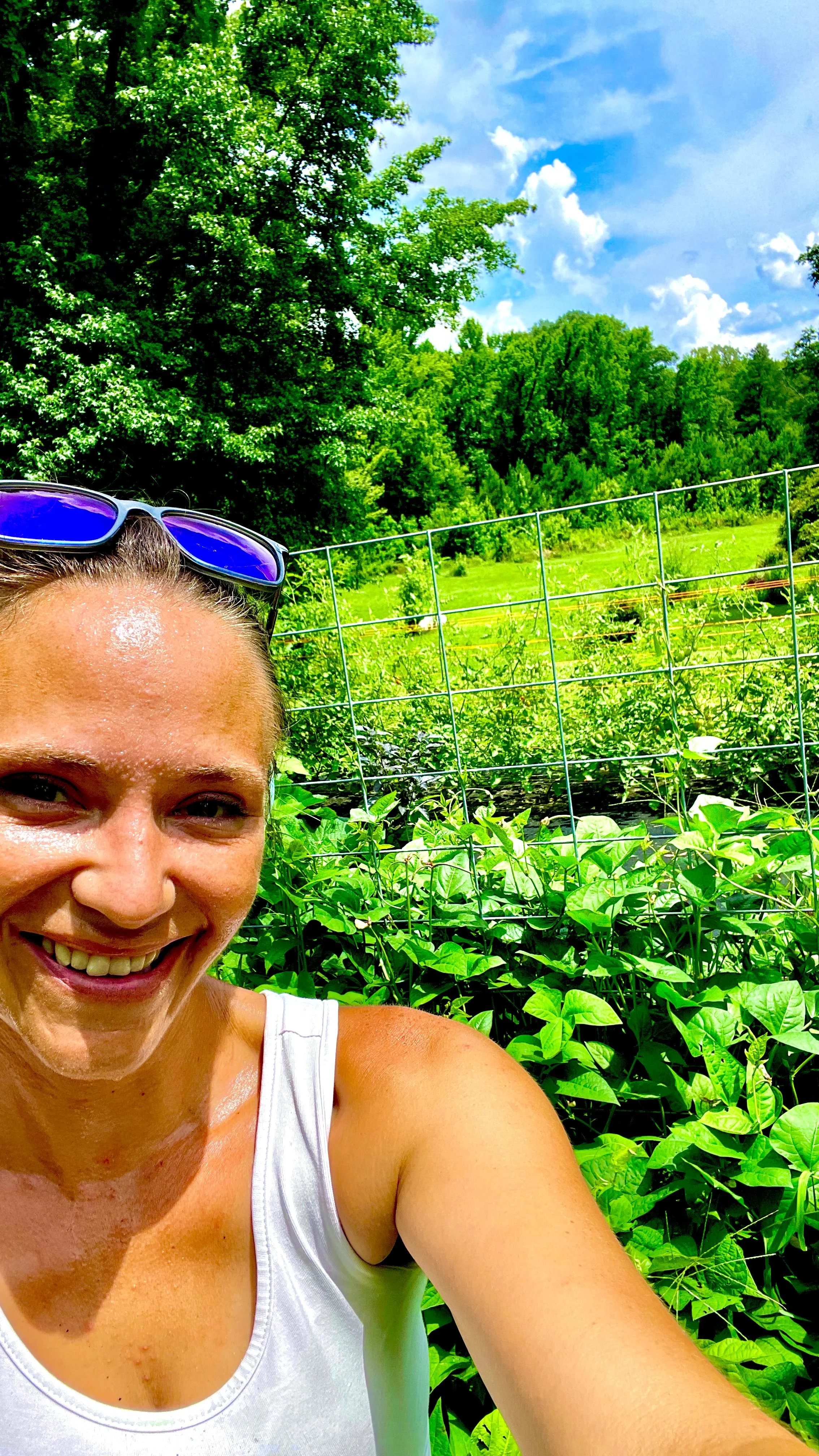 Close-up of a smiling woman with sunglasses on her head, wearing a white tank top, in a lush garden with green plants and a metal fence, under a partly cloudy sky.