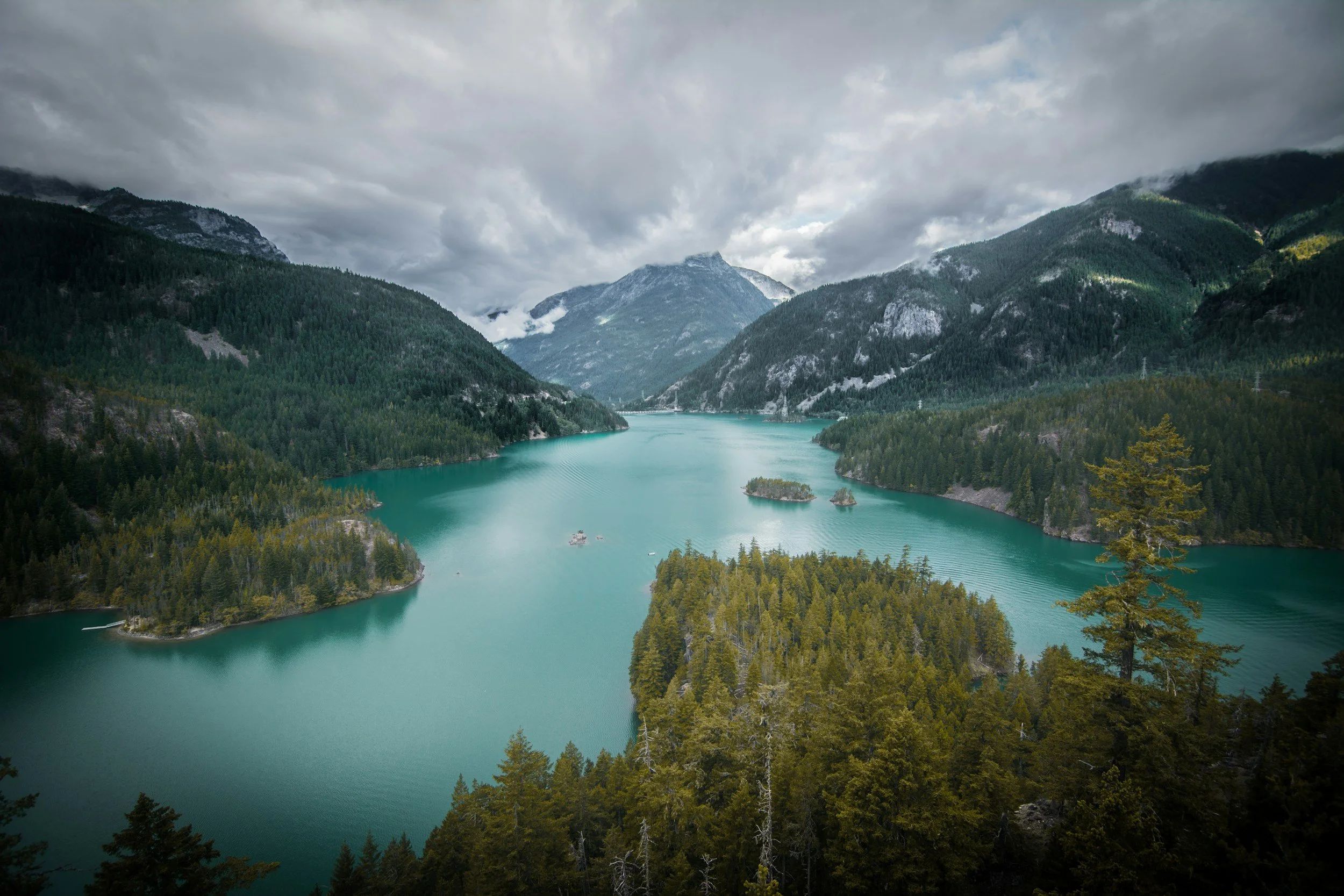 A scenic view of a mountain range with a lake in the foreground, surrounded by pine trees and a cloudy sky.