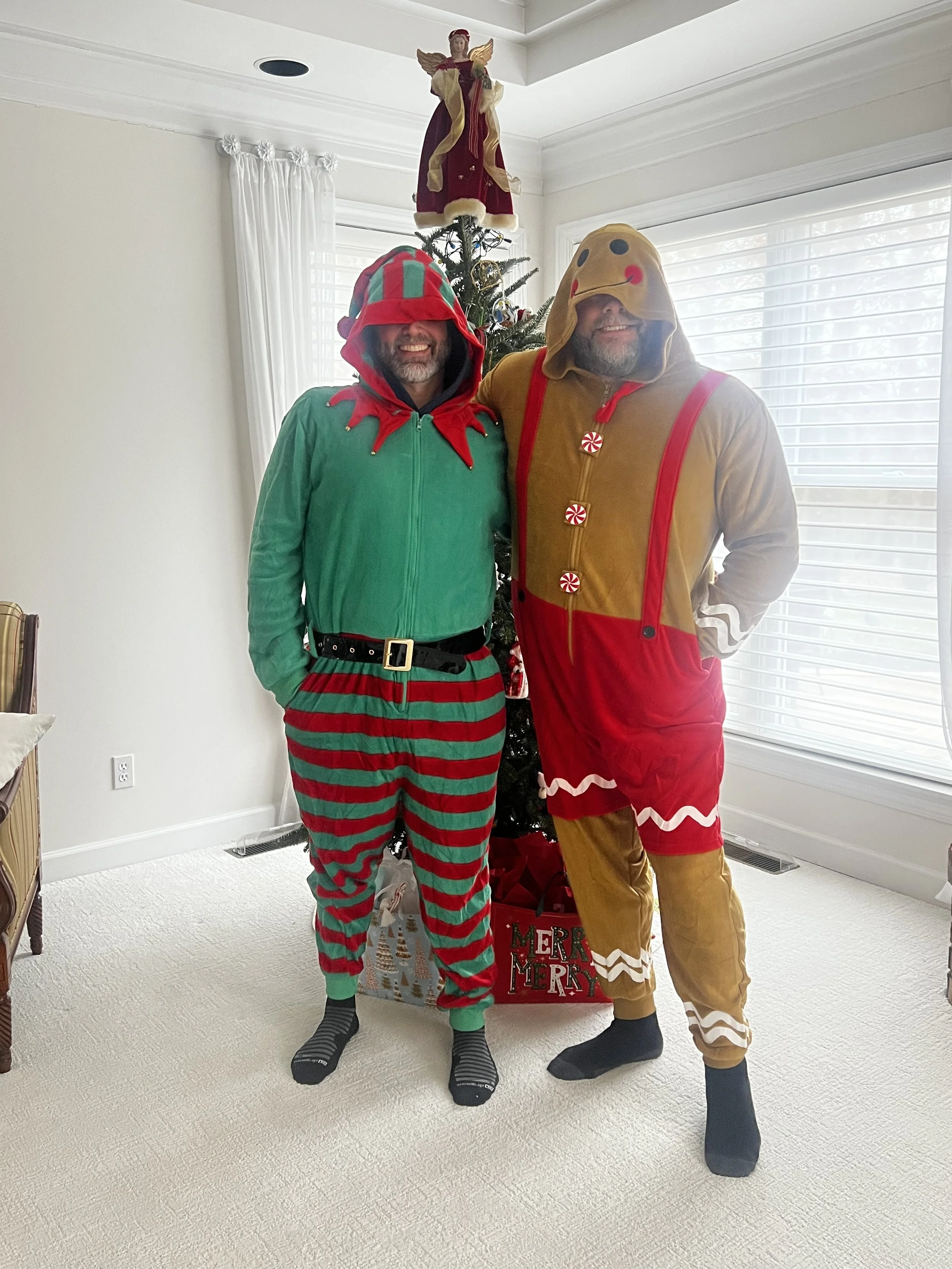 Two men in Christmas pajamas and costumes, one dressed as an elf and the other as a gingerbread person, standing in front of a decorated Christmas tree and a window with blinds.