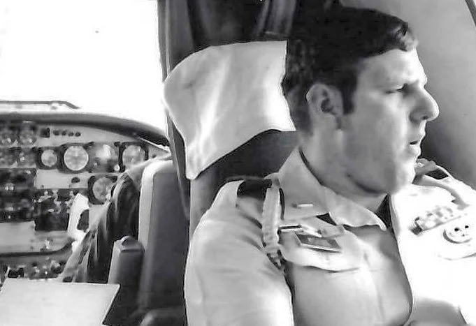 Black and white photo of a man in a military uniform sitting in a cockpit, looking out the window with a serious expression, with aircraft instruments visible behind him.