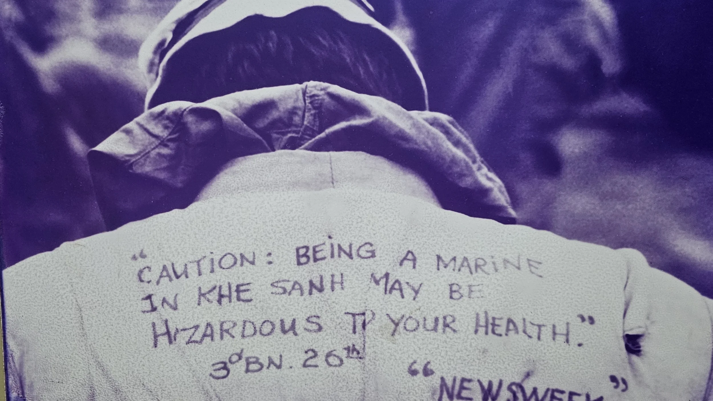 A person with long hair wearing a beanie, holding a piece of paper with handwritten text. The paper contains a quote about being a marine, health, and hazards, with a date at the bottom.
