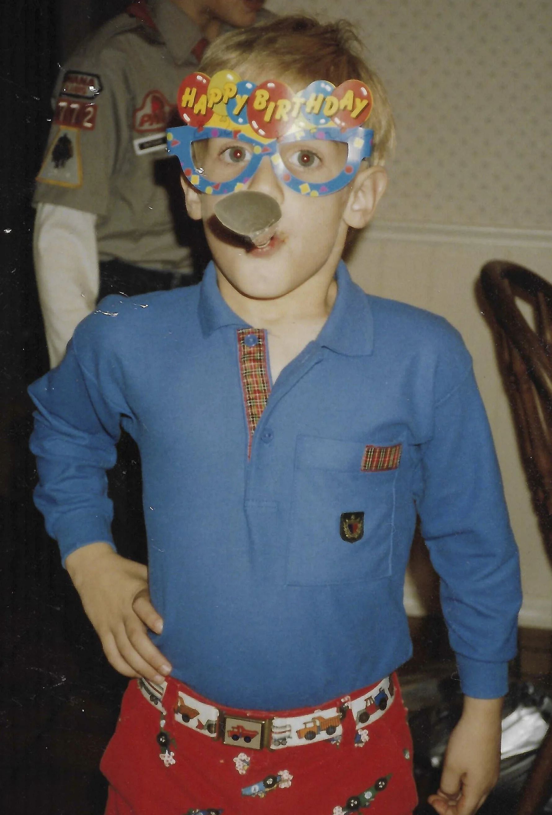 A young boy wearing holiday-themed sunglasses with the words 'Happy Birthday' on top, a blue shirt, red shorts with vehicle patterns, and decorated with various crafts and puzzle pieces. An adult dressed in a uniform stands in the background.