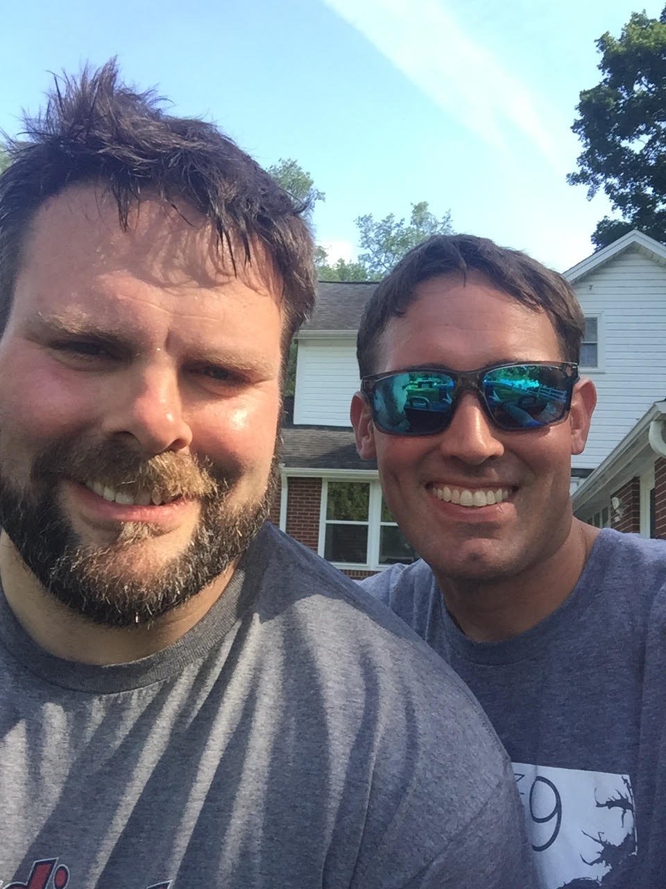 Two smiling men taking a selfie outdoors in front of a house, one wearing sunglasses and the other with wet hair and a beard.