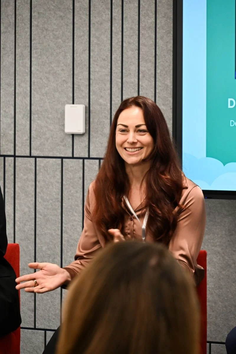 Smiling woman with long red hair speaking at an indoor event, seated near a gray wall with vertical black lines and a large screen in the background.