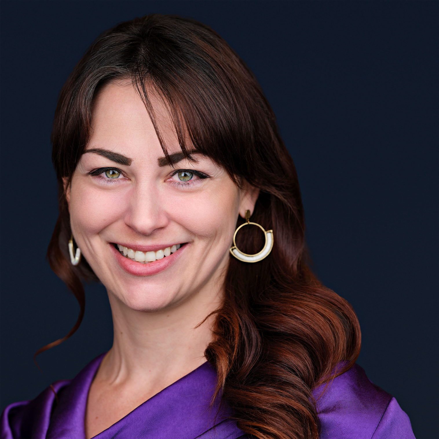 Close-up portrait of a woman with brown hair, green eyes, gold earrings, smiling, wearing a purple garment, against a dark background.