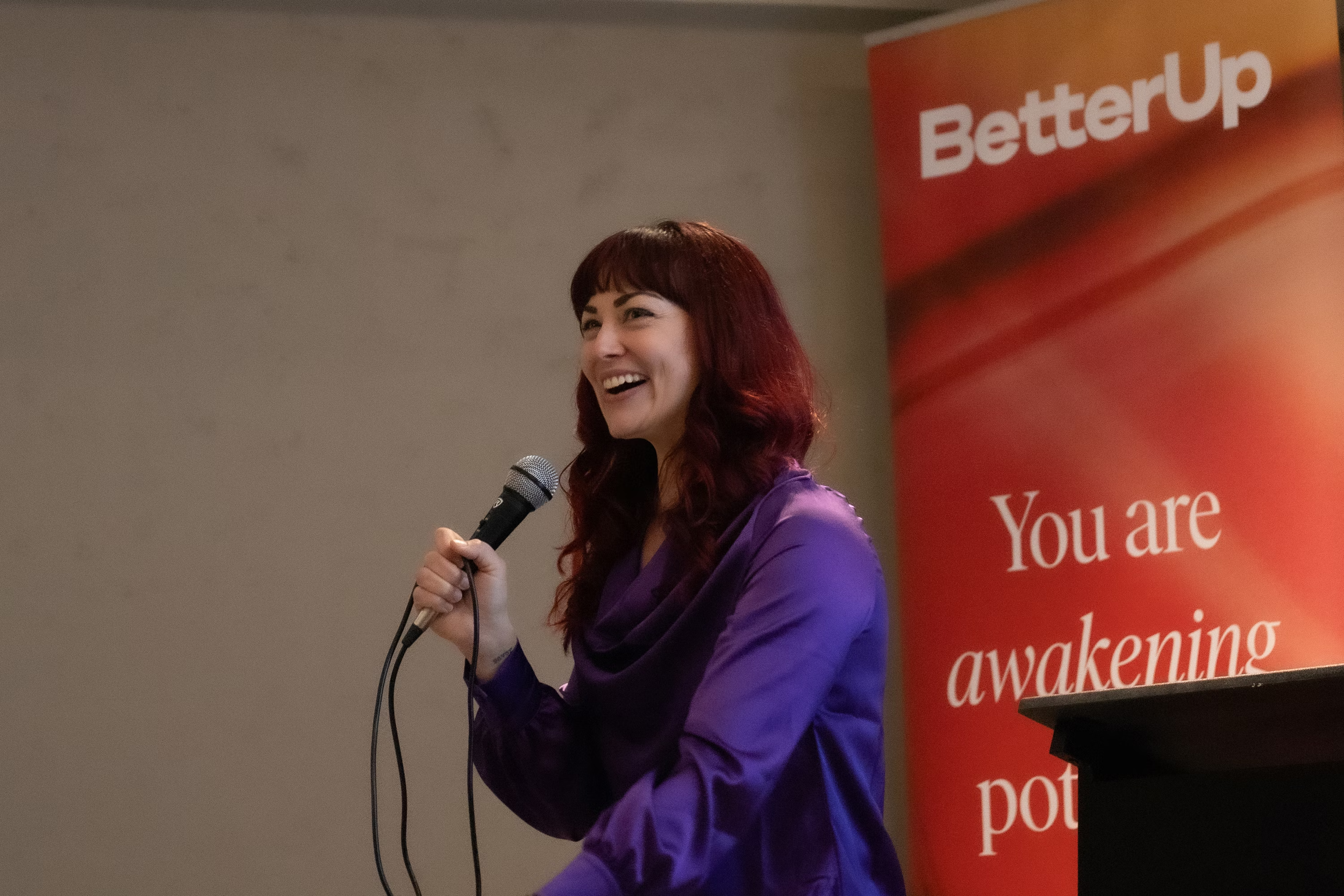 A woman with red hair speaking into a microphone at an event, standing in front of a red and orange banner that reads 'Better Up You are awakening'.
