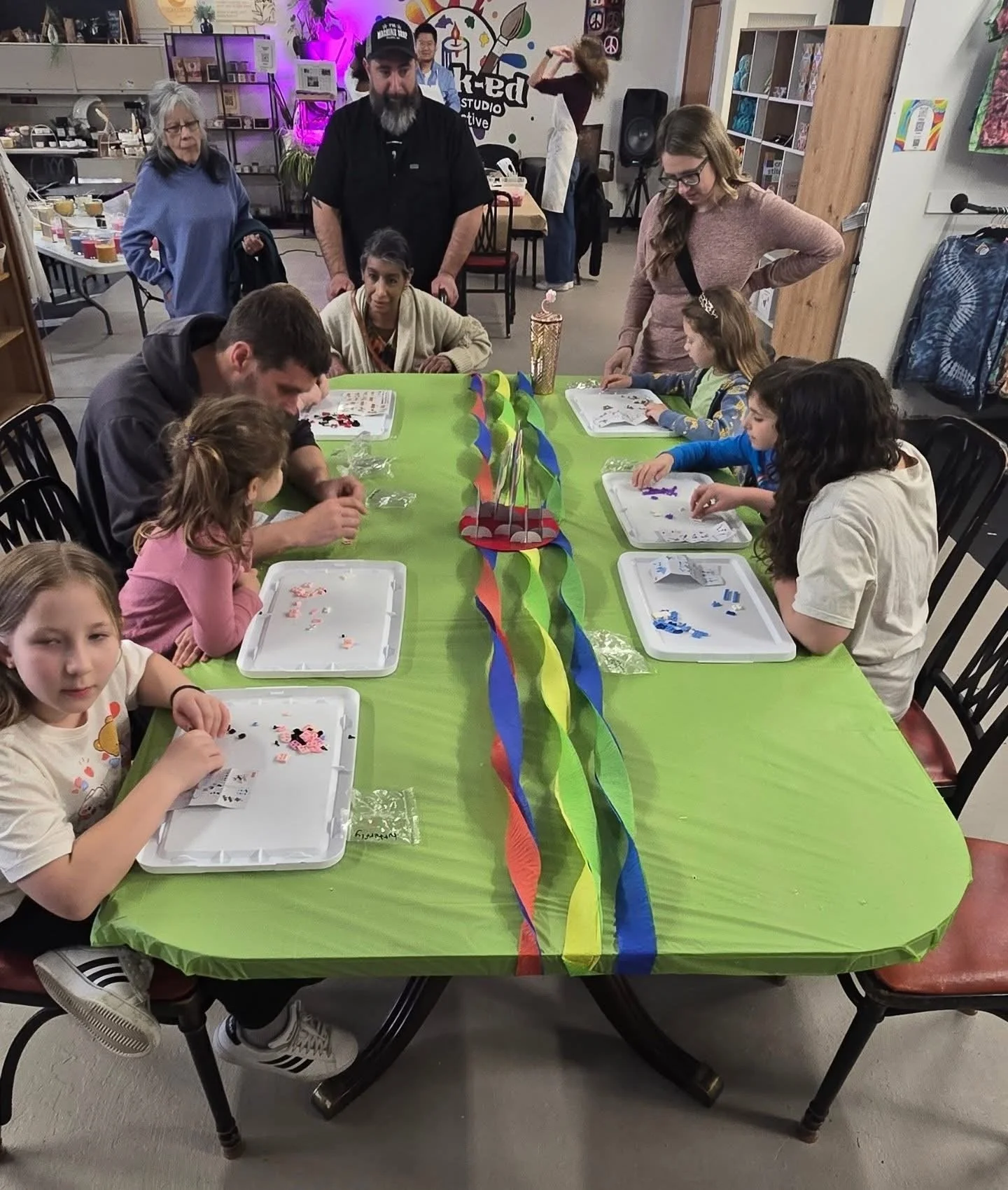 Children and adults gathered around a decorated table with colorful streamers, engaging in crafting activities with small tiles and stickers in a community or art studio setting.