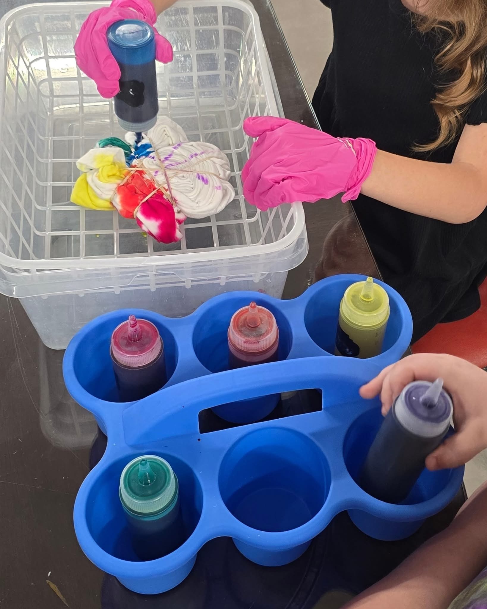 A person wearing pink gloves is filling squeeze bottles with colored liquid dye, used for tie-dyeing fabric. There are multiple squeeze bottles with different colors placed in a blue plastic holder, and the person is dyeing a white shirt in a plastic
