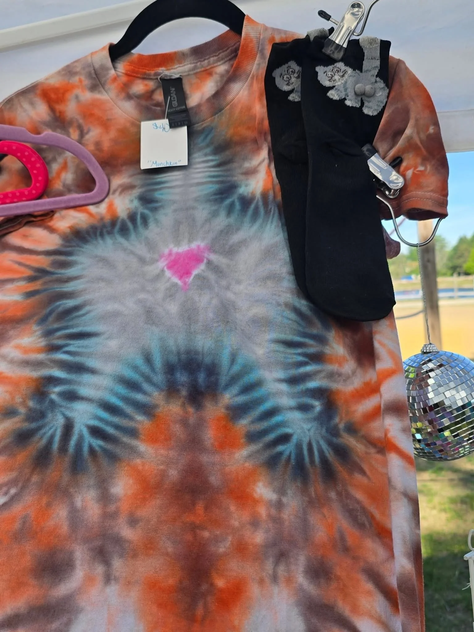 A tie-dye T-shirt with a multicolored pattern, hanging on a hanger. A black sock with a floral pattern and buttons is clipped to the hanger. A small disco ball ornament hangs in the bottom right corner.