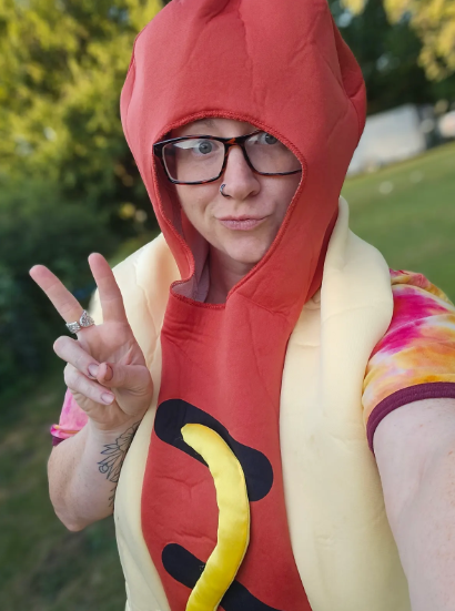 Person outdoors dressed as a hot dog, wearing a red hood, yellow costume with mustard and ketchup designs, and glasses, making a peace sign.