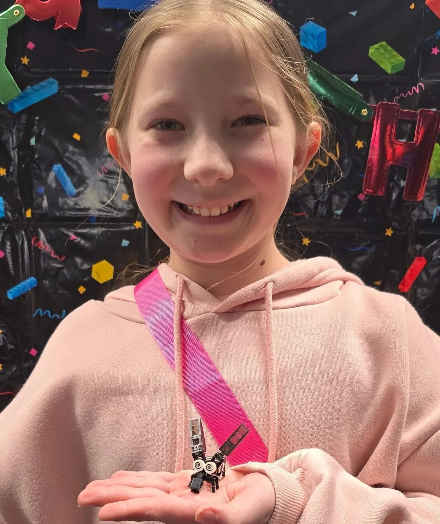 A young girl with blonde hair and a pink hoodie holding a small black and white LEGO dog in her palm, standing in front of a colorful background with stars and confetti.
