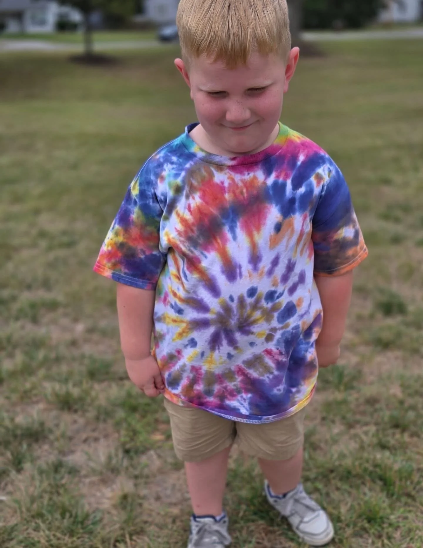 A young boy with red hair wearing a colorful tie-dye t-shirt, beige shorts, and sneakers standing outdoors on grass.