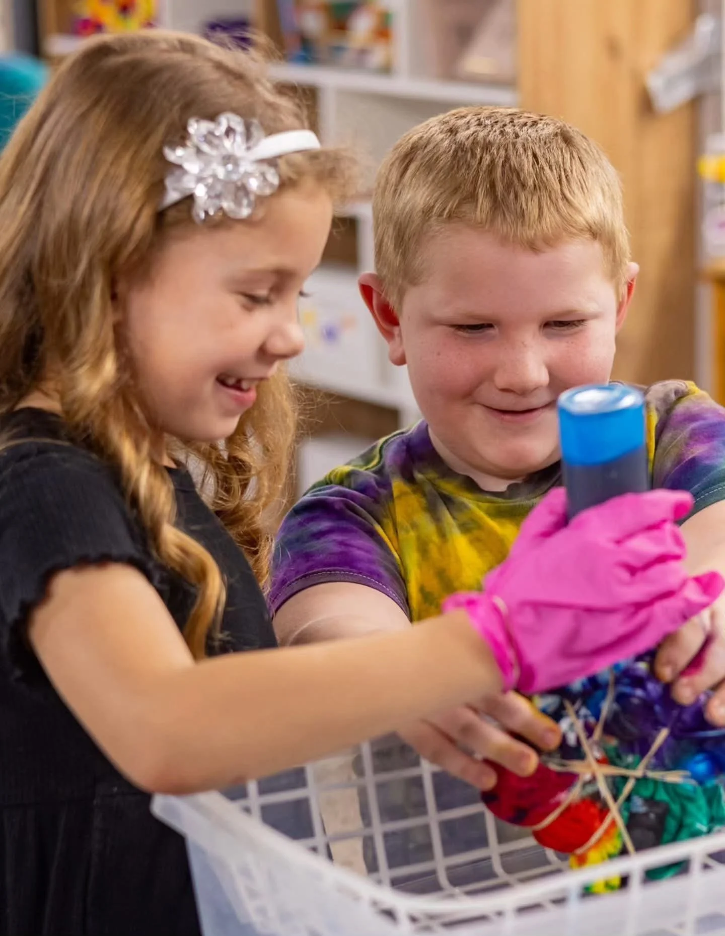 Two children, a girl with red hair and a boy with red hair, are engaging in a science activity. The girl is smiling and looking at the boy, who is holding a test tube or small container with colorful strands of yarn, wearing pink gloves. They are in 