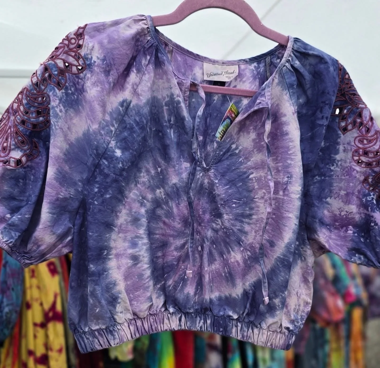 Purple tie-dye blouse with lace-up detailing on the shoulders, hanging on a pink hanger in a clothing store.